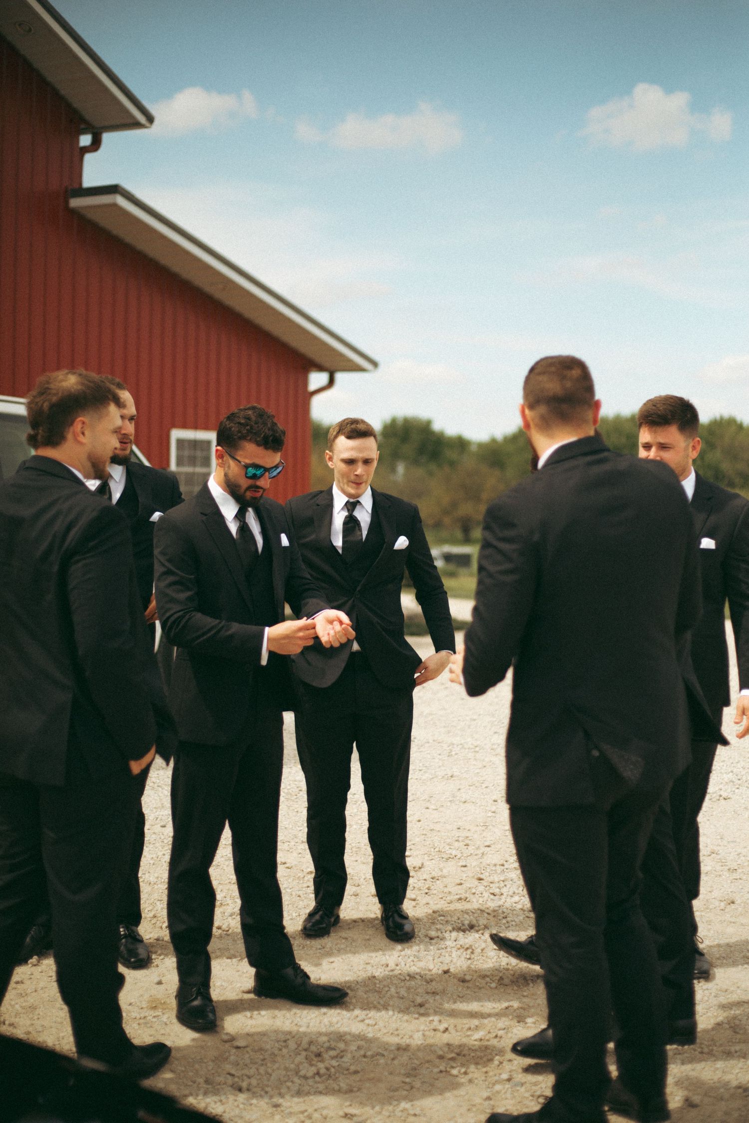 Groomsmen in black suits gather outside red barn for pre-wedding conversation.