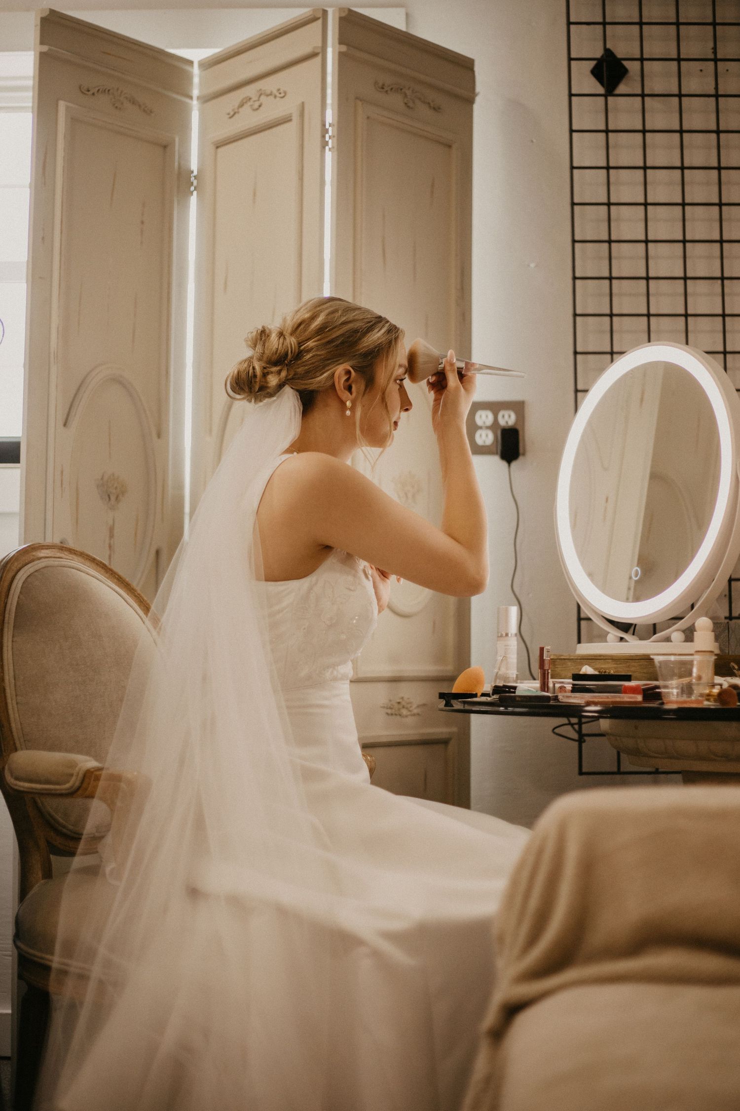 Figure in white wedding dress applies makeup in front of vanity mirror.