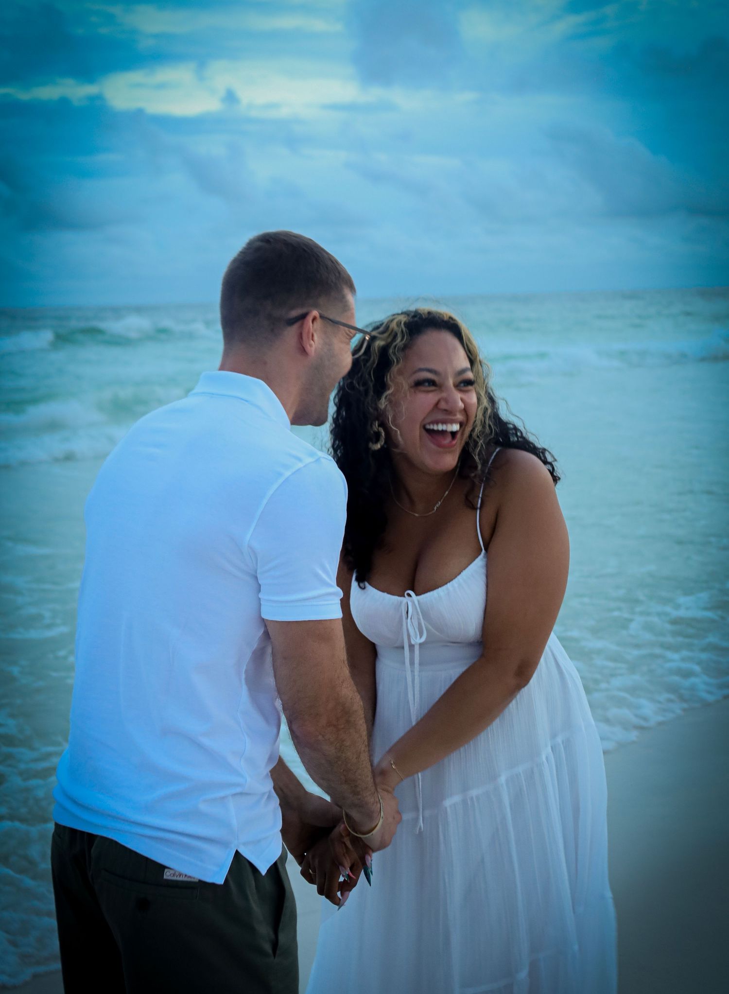 A couple in white clothing shares joyful moments during a beach photoshoot with turquoise waters in the background.