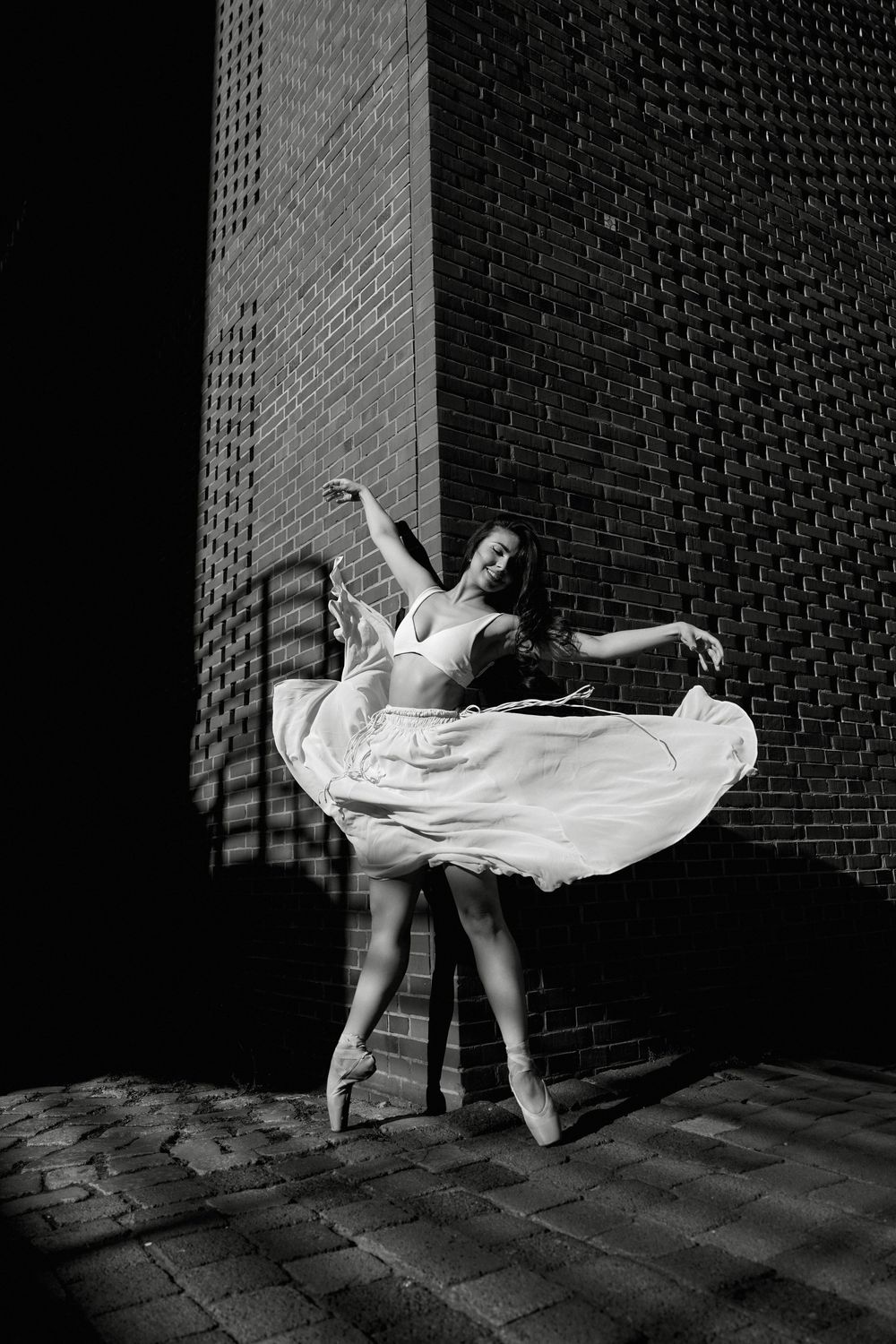 Figure in flowing white dress twirling against brick wall in black and white.