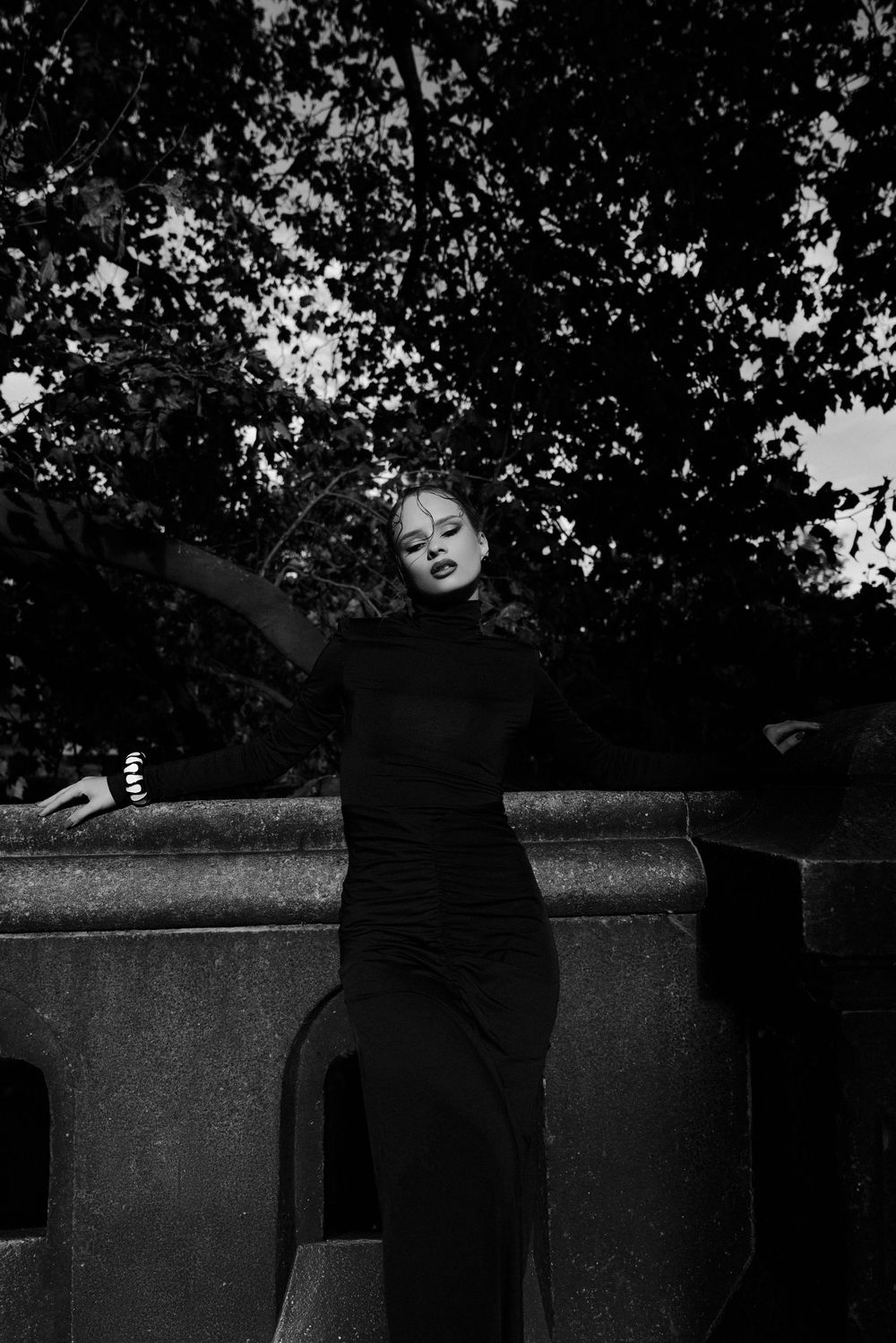 Artistic black and white photo of a person reclining on stone wall under tree branches.