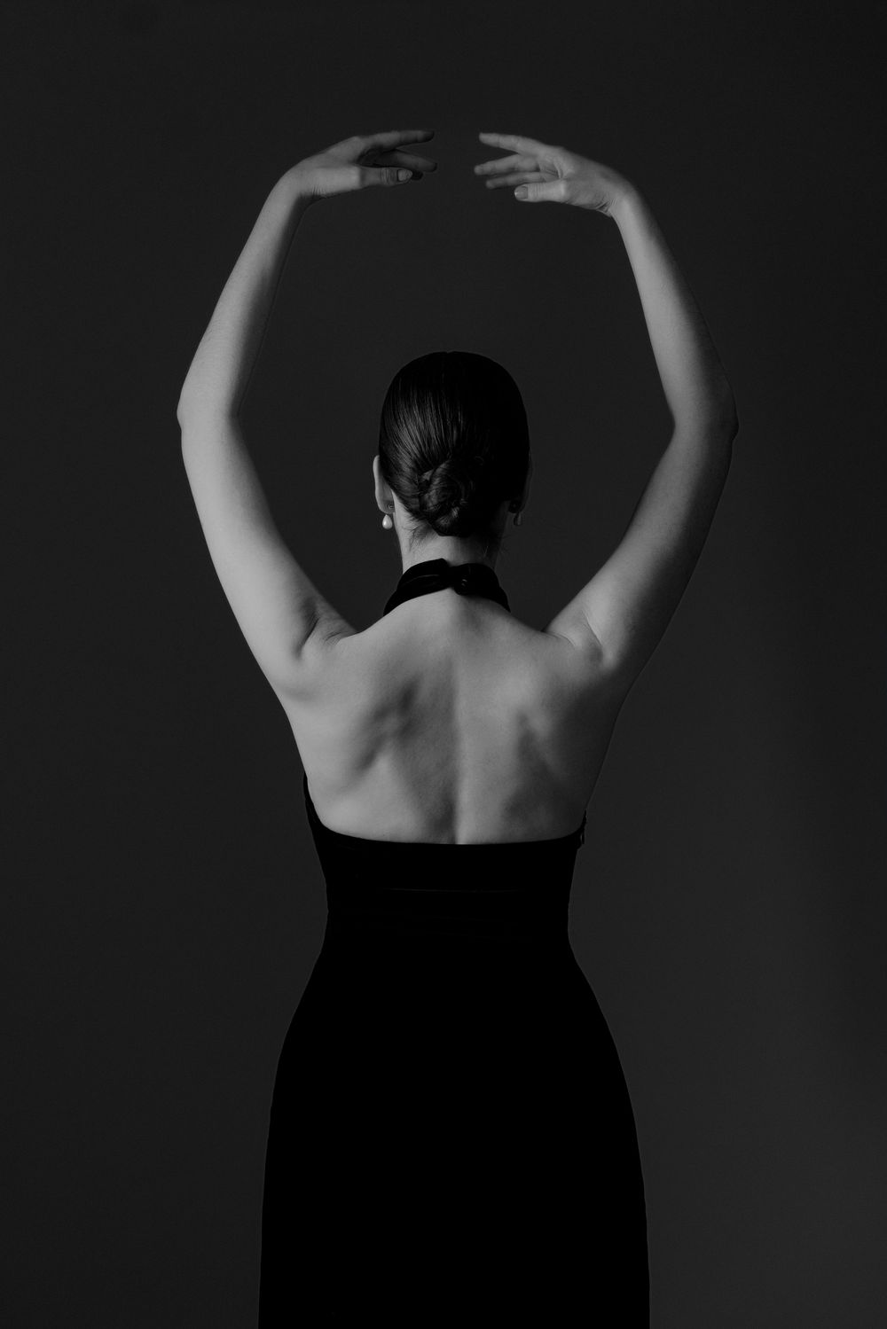 Black and white photo of a woman in a backless black halter dress with arms raised gracefully overhead.