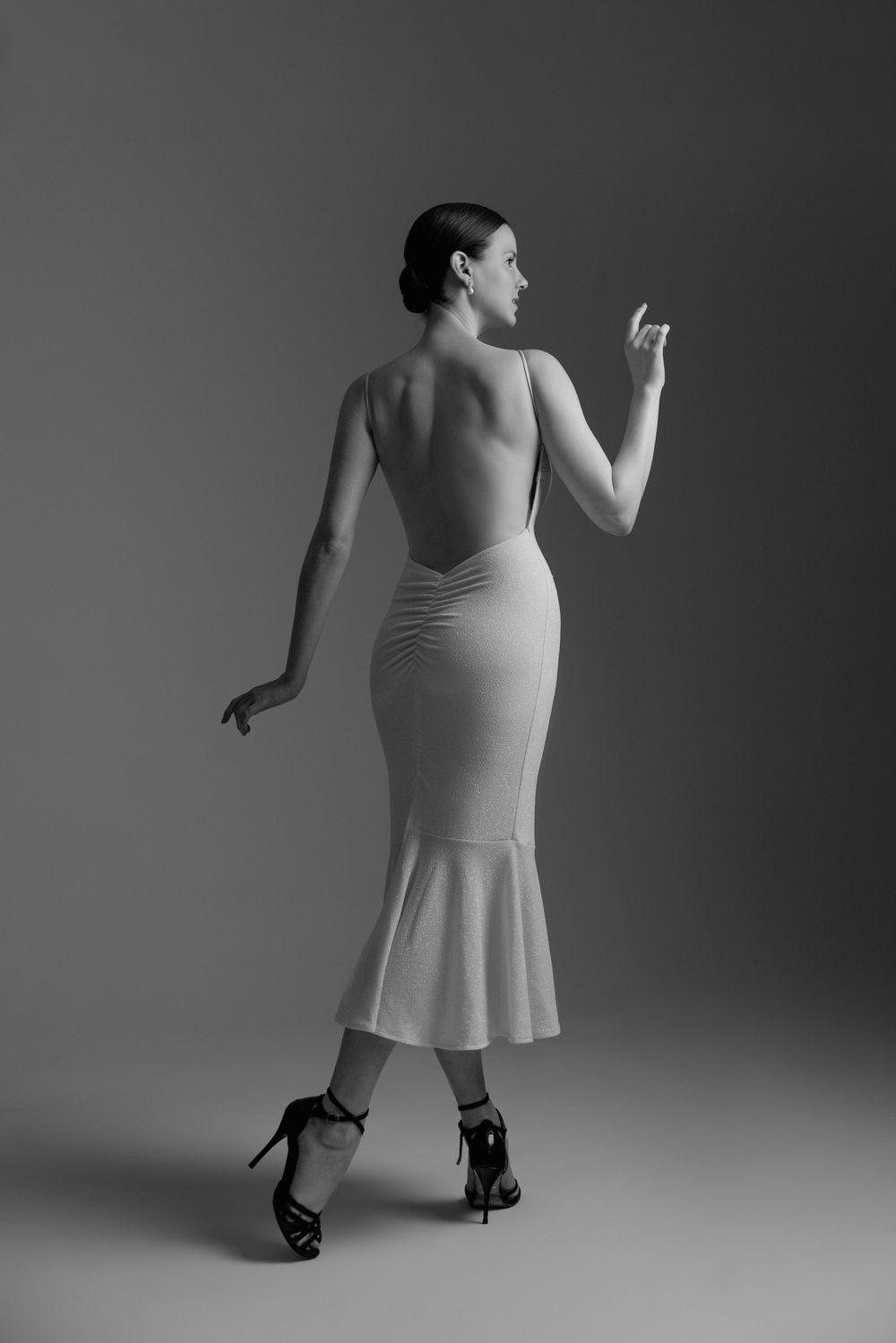 Black and white photo of a woman in a backless midi dress and heels, posed elegantly in a studio setting.