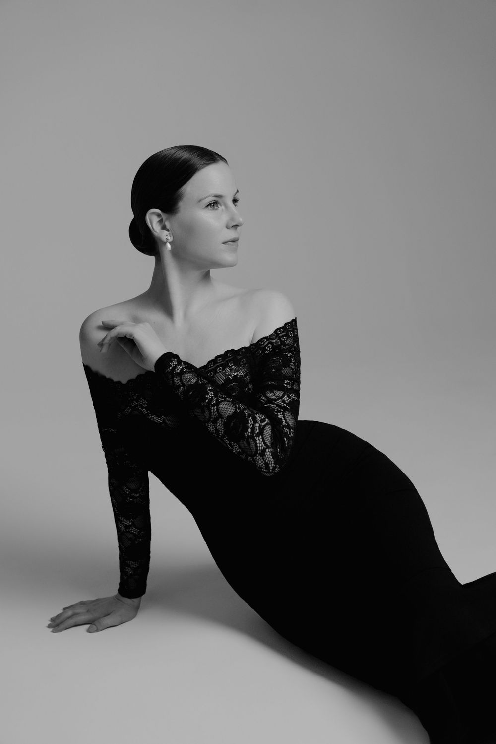 Elegant woman in black lace off-shoulder gown posing gracefully on floor in black and white studio portrait.