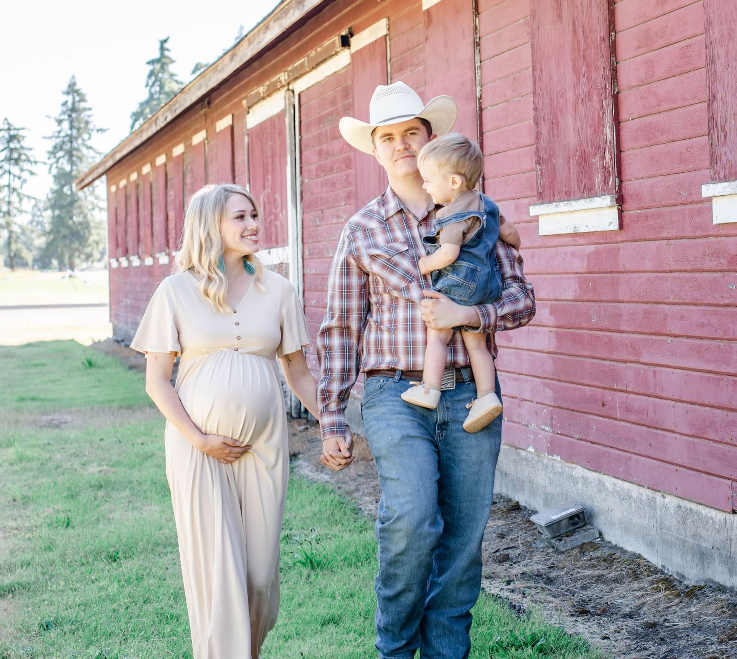 Couple posing for maternity photos against weathered red barn in countryside setting.