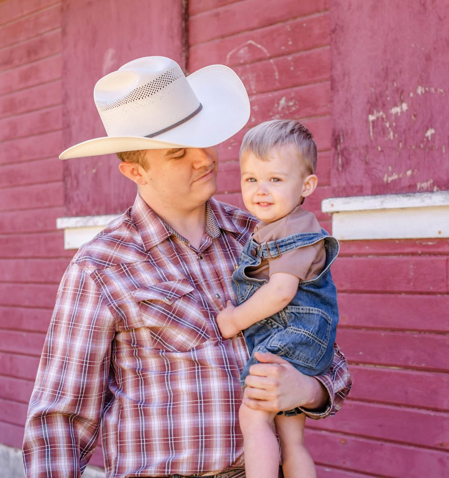 A parent in plaid shirt and cowboy hat holding small child in overalls against red barn wall.