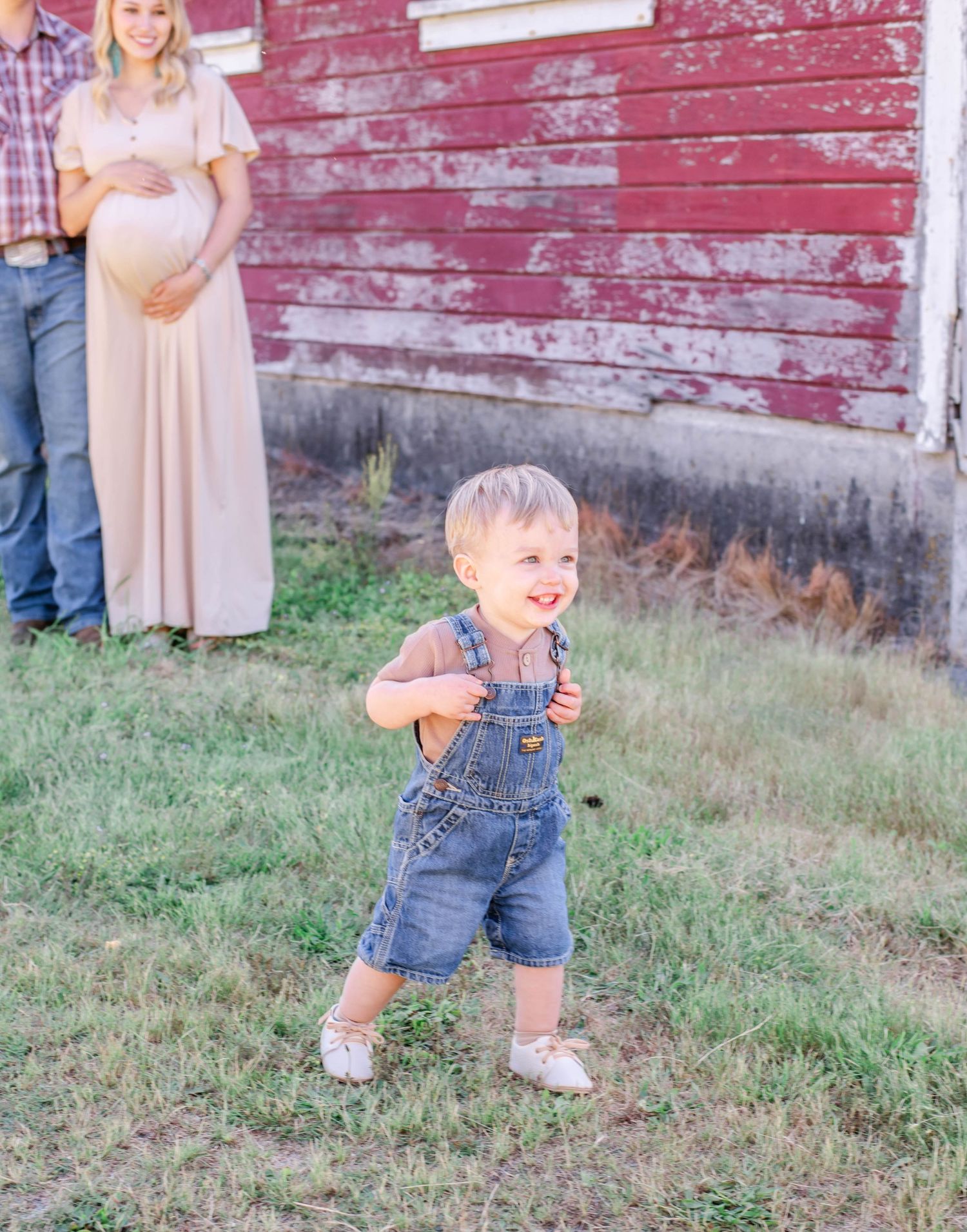 Toddler in denim overalls playing on grass near red barn wall.