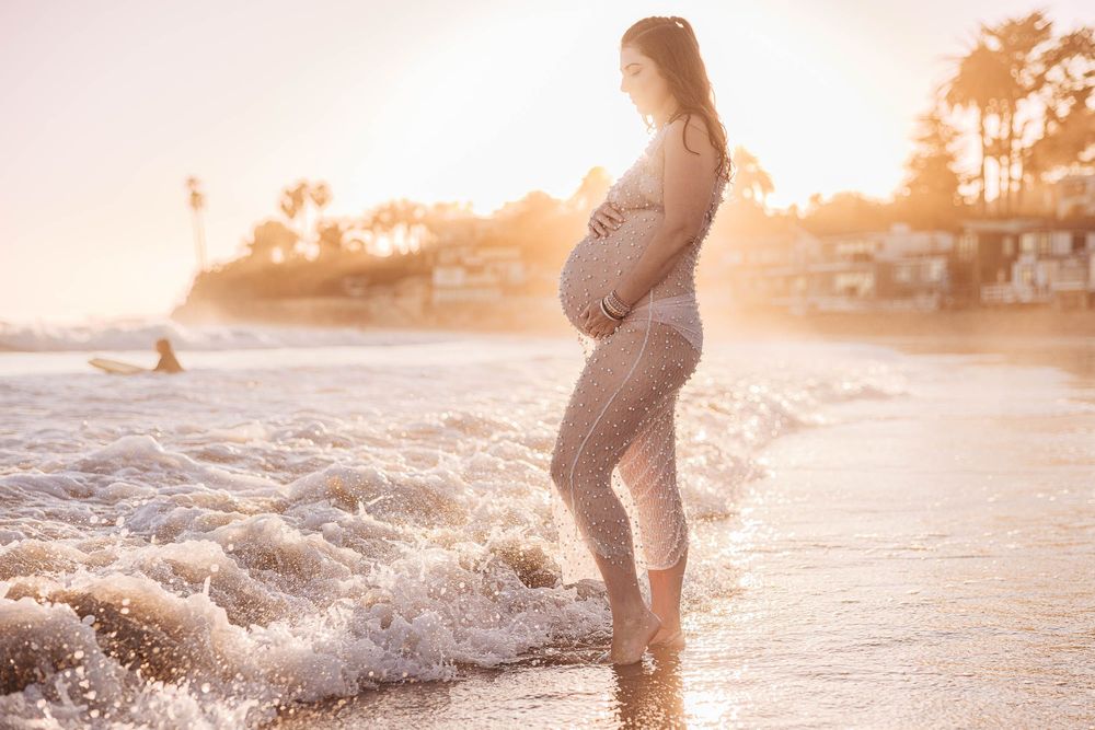 A silhouetted pregnant figure splashes in ocean waves at sunset creating a magical beach maternity scene.