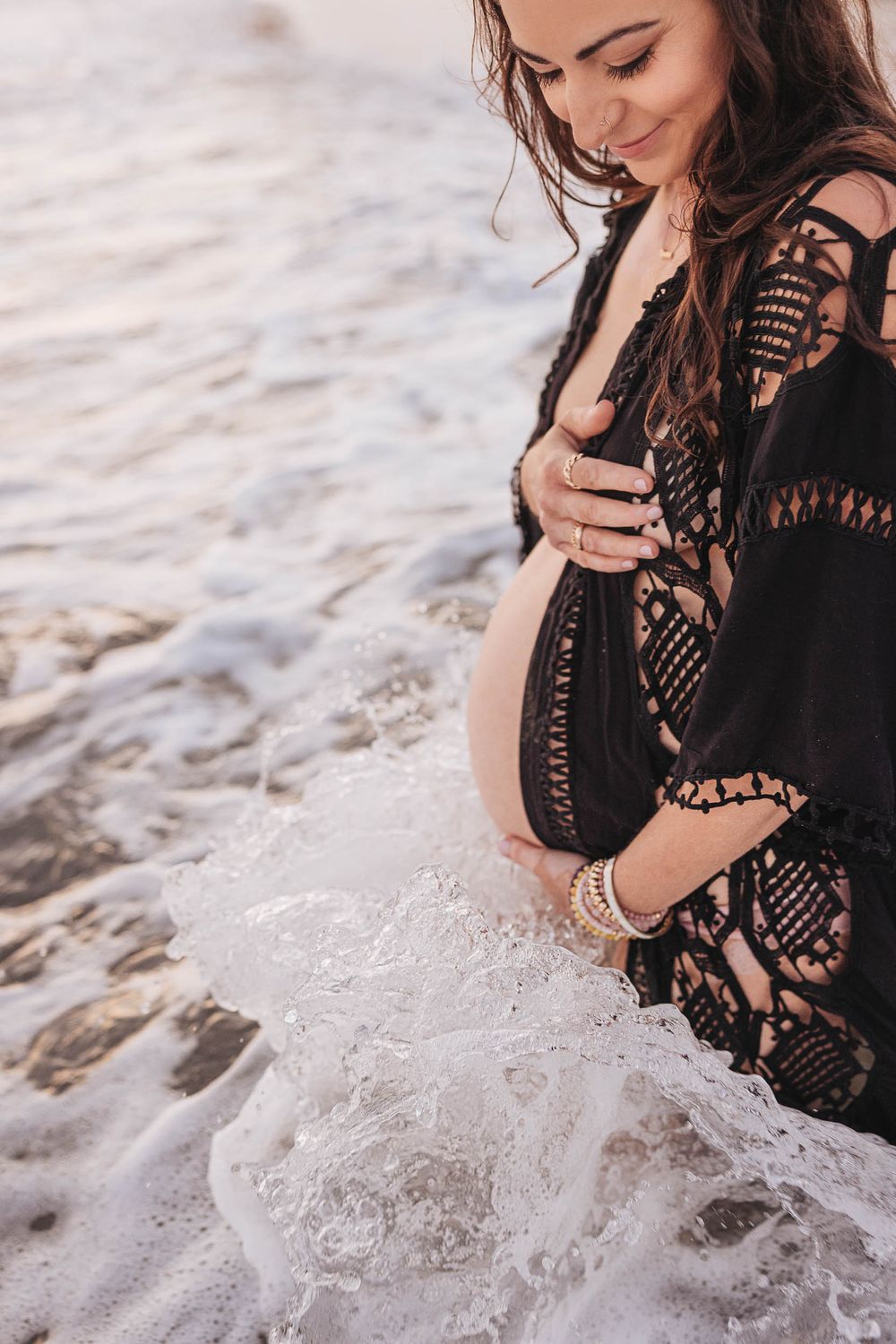 A pregnant woman wearing a black crochet cover-up stands at the beach while ocean waves gently wash over her feet.
