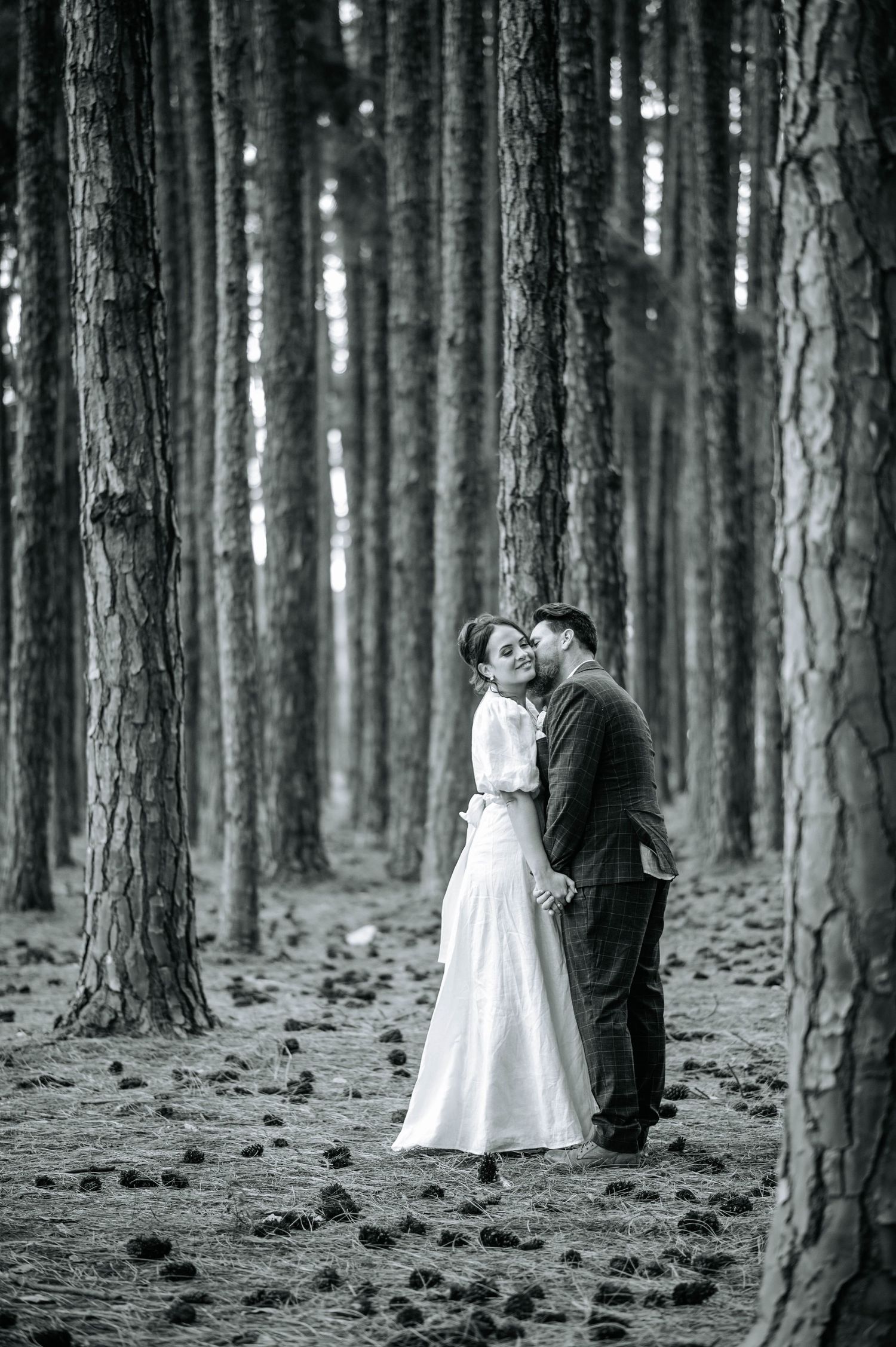 A black and white portrait of newlyweds embracing in a pine forest.