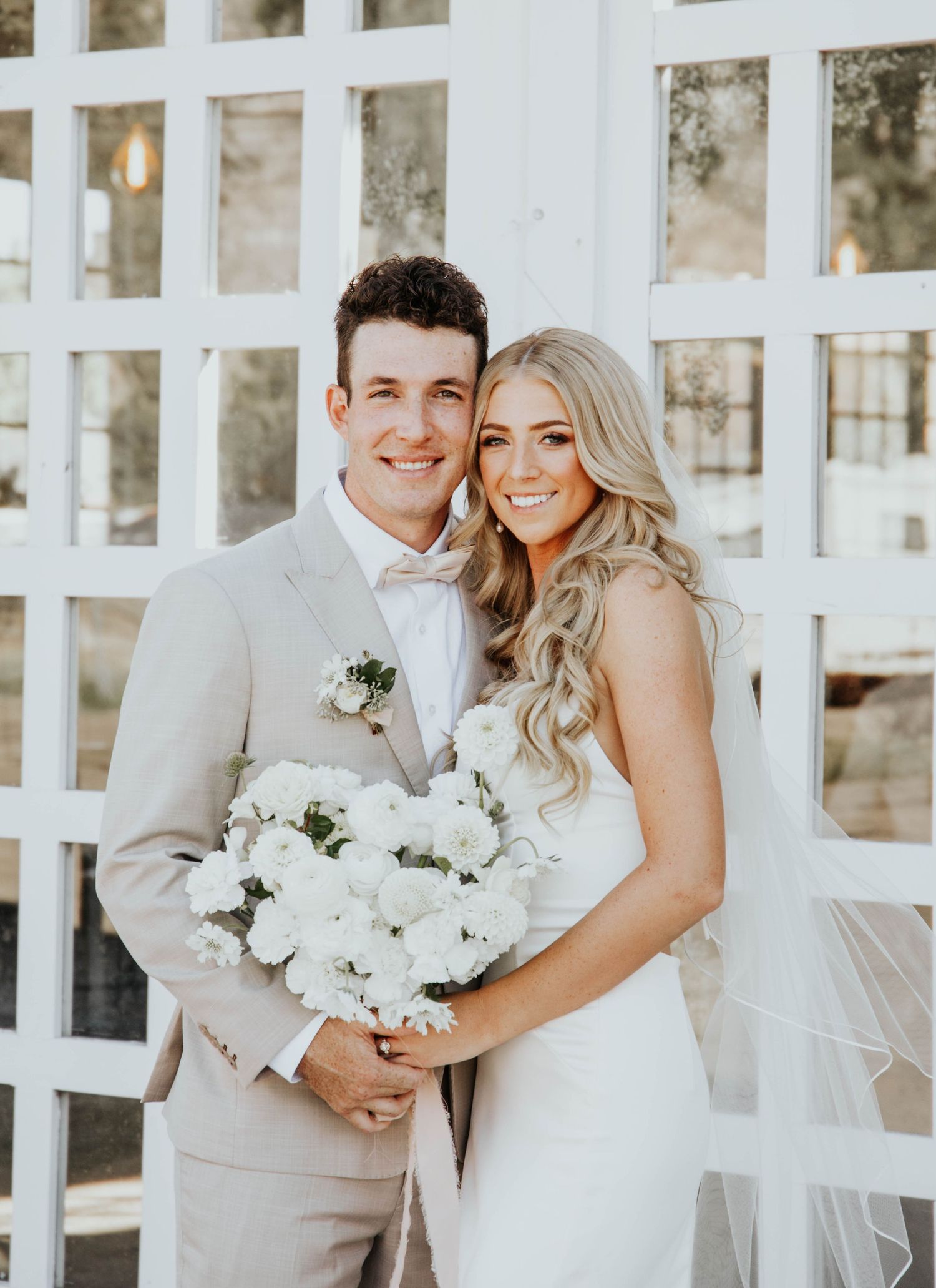 Wedding couple poses against white lattice backdrop with white floral bouquet.