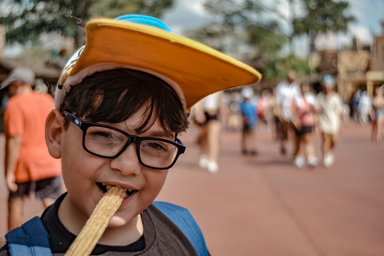 Young tourist wearing glasses and orange hat enjoys a snack at a crowded theme park.
