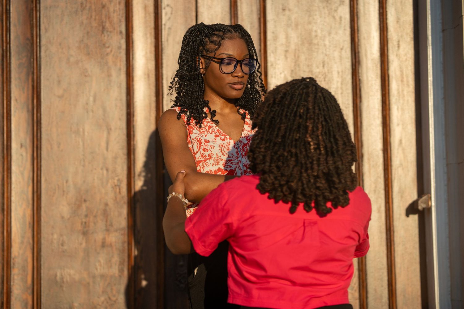 A woman in glasses and a coral top stands against a wooden wall, facing a person with long braids in a red shirt in warm sunlight.