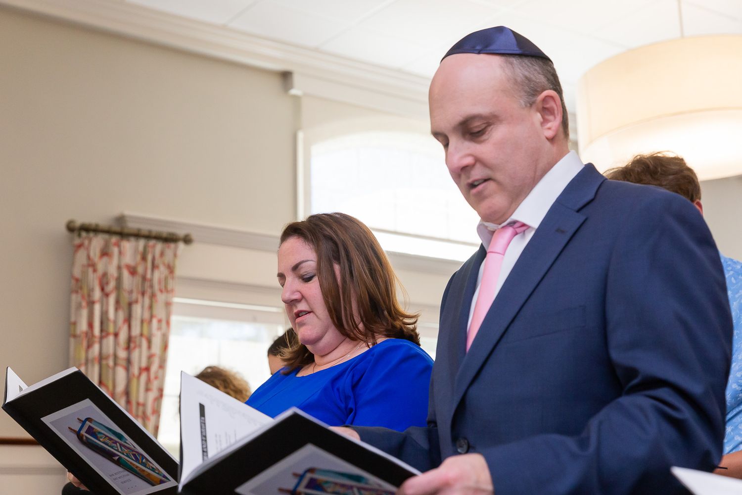 Two people reading from prayer books during a religious service while wearing formal attire.