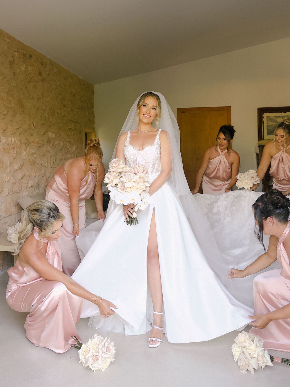 Bridesmaids in blush pink dresses help arrange the bride's elegant white wedding gown and long veil in preparation room.