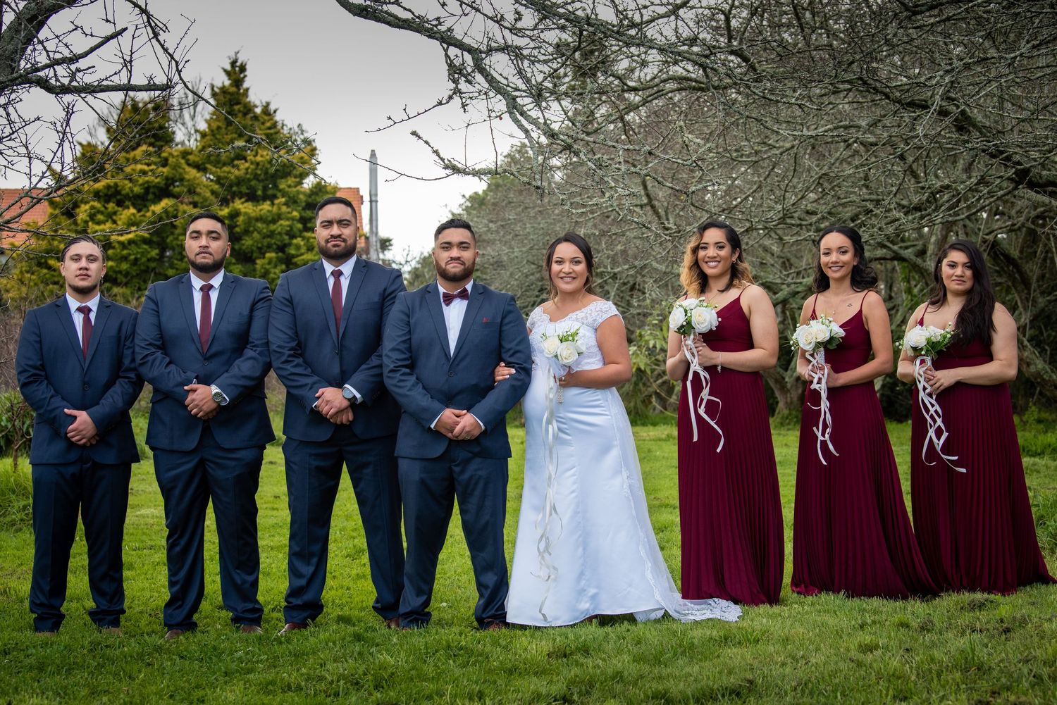 Wedding party in navy suits and burgundy dresses posing outdoors under blooming trees.