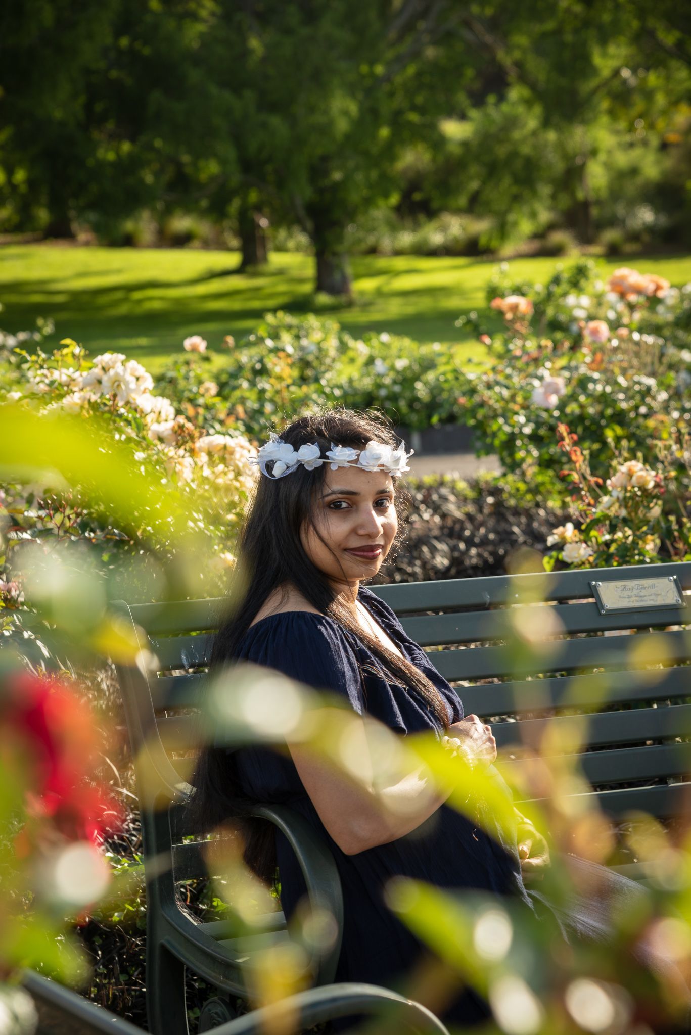 Figure wearing floral crown sits on bench in sunny garden with blooming roses.