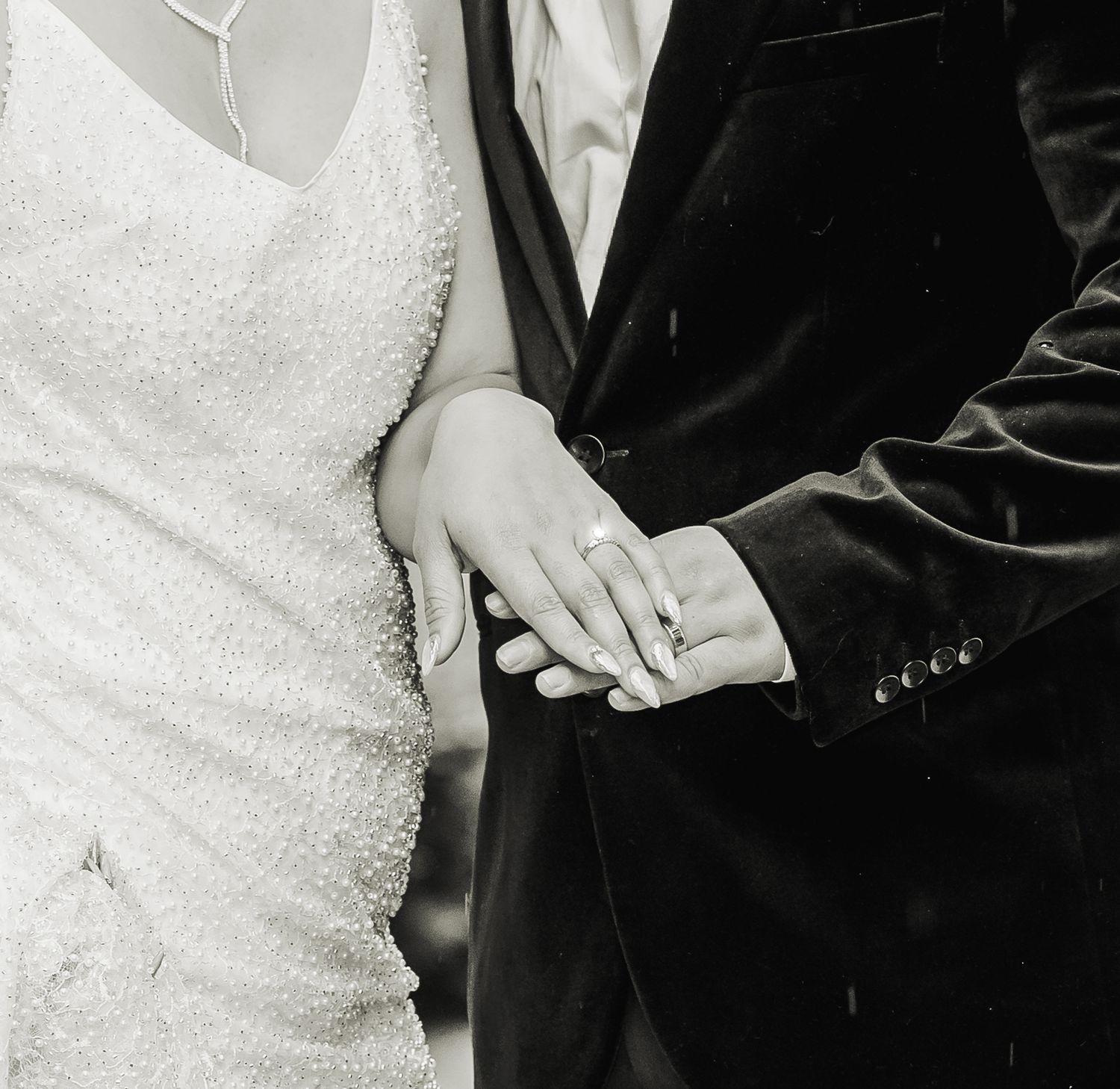 Black and white close-up photo of newlyweds showing hands wearing wedding rings with dress and suit in view.