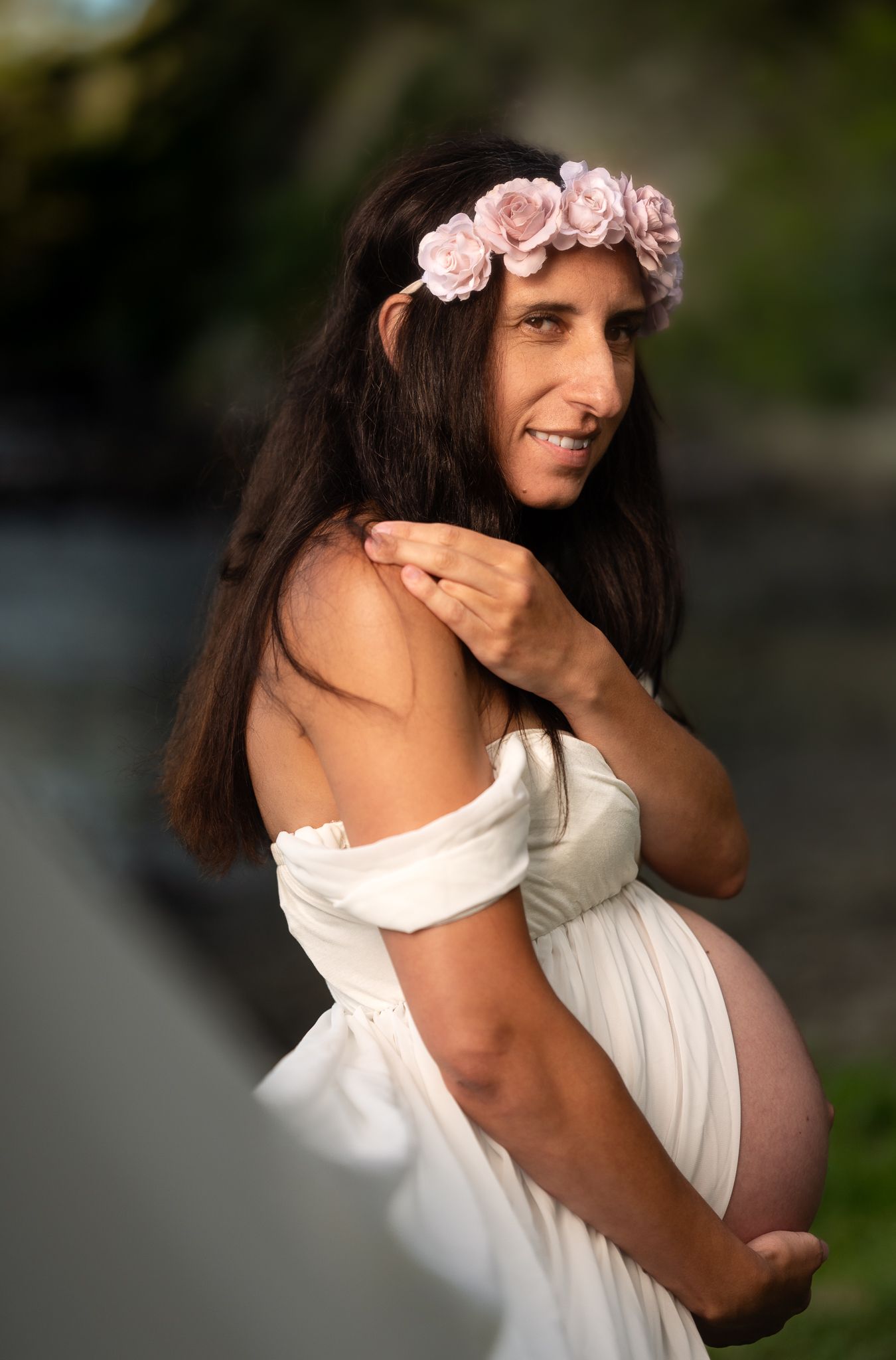 Beautiful maternity portrait outdoors with flowing white dress and delicate pink flower crown in natural lighting.