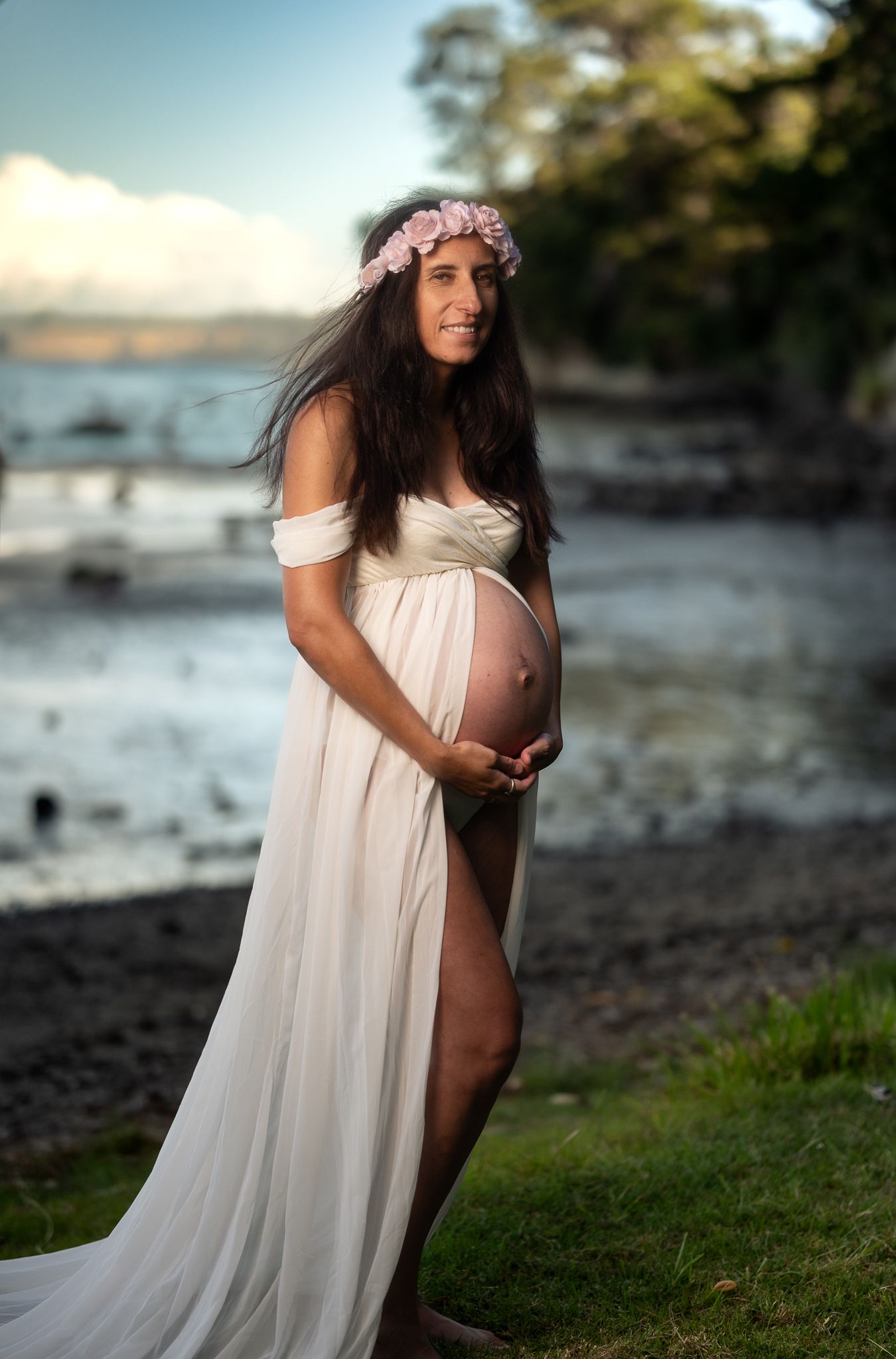 Maternity photo on a beach at sunset features a flowing white dress and floral crown against an oceanic backdrop.