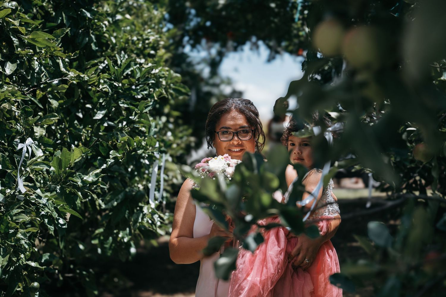Through green leaves and branches, people stand together outdoors on a sunny summer day.
