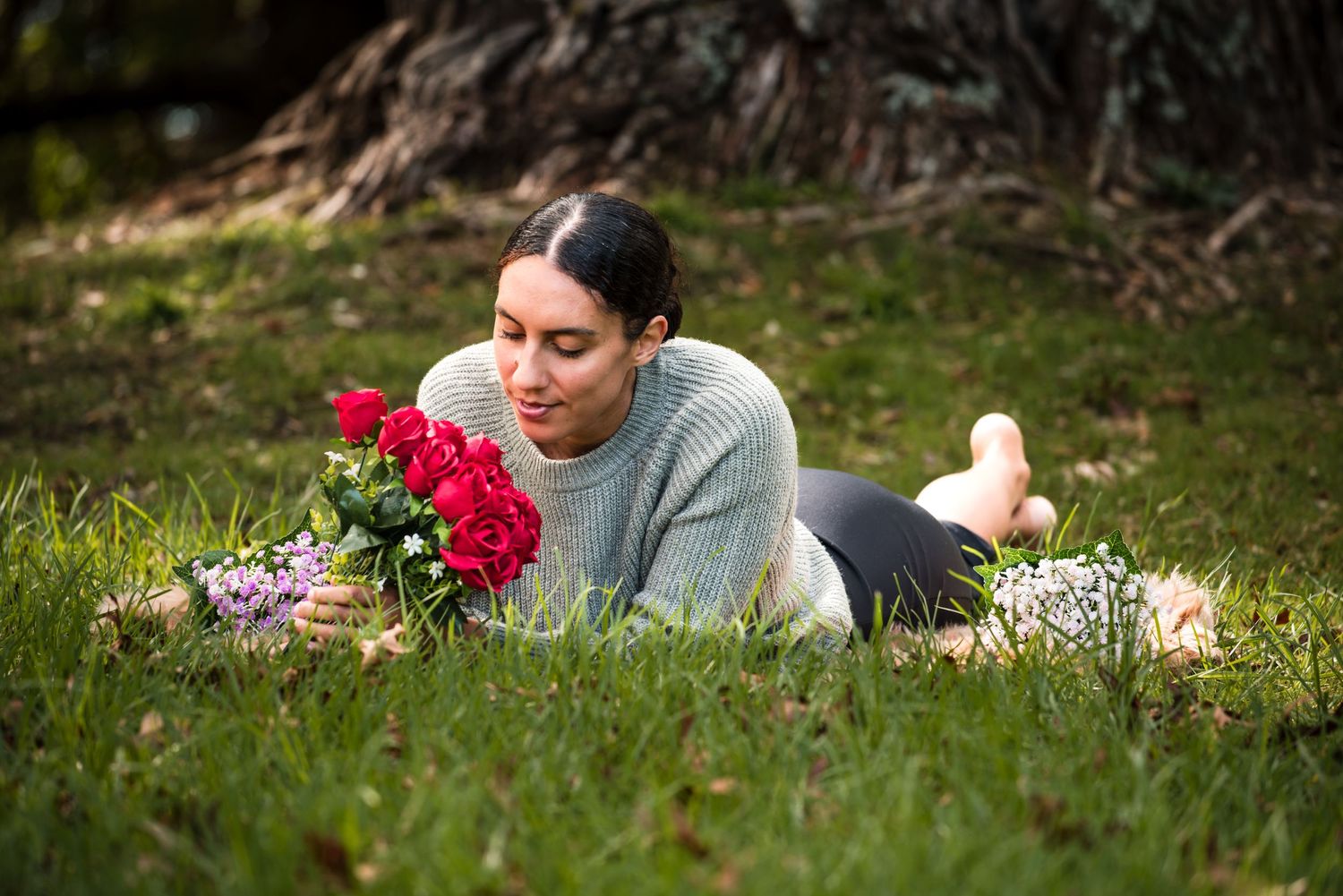 Someone lying on grass smelling vibrant red roses in a soft gray sweater.