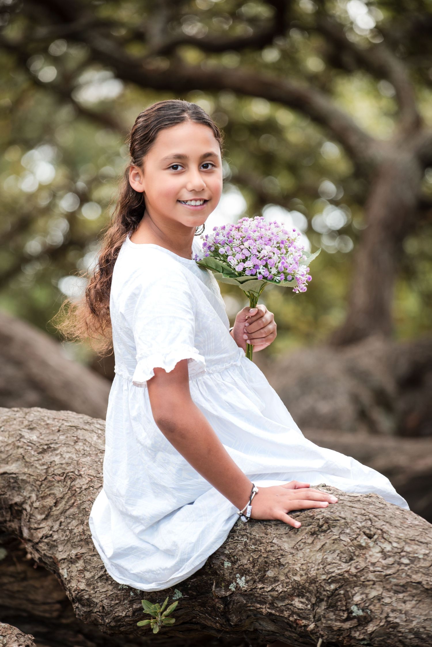 Girl in white dress sitting on tree branch holding purple flowers in outdoor portrait.
