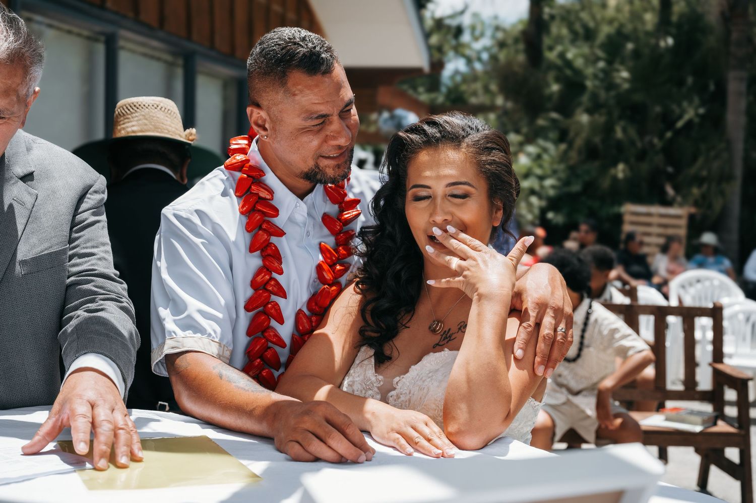 Wedding couple sign marriage documents together at outdoor ceremony with laughing smiles and red lei decorations.
