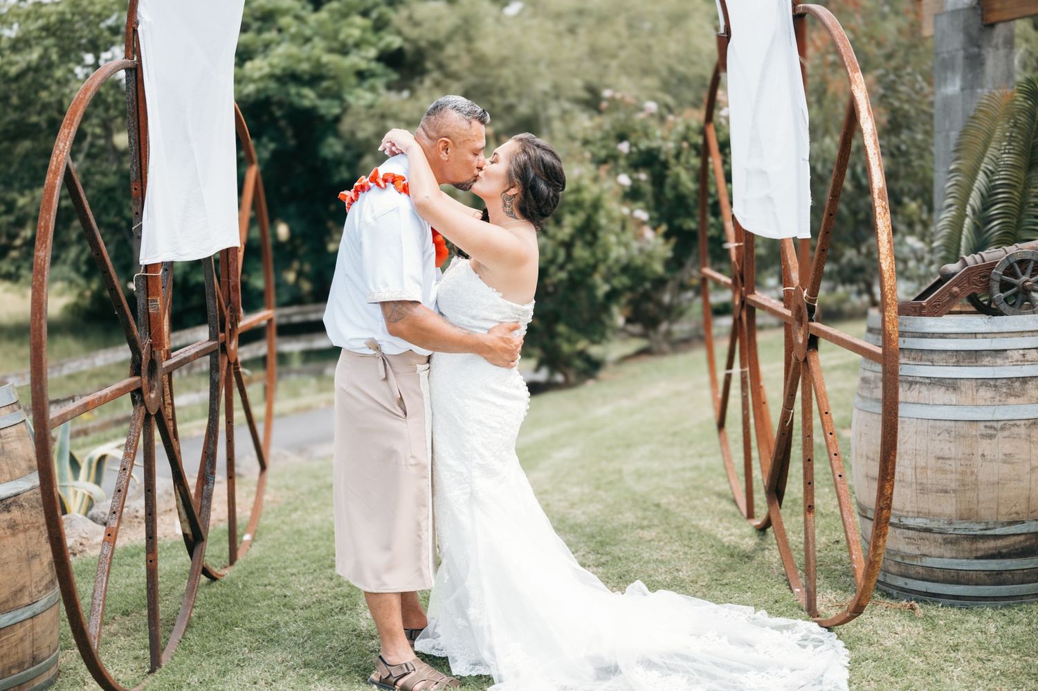 A romantic outdoor wedding ceremony with white fabric draped on rustic metal arches and wooden barrels in a garden setting.