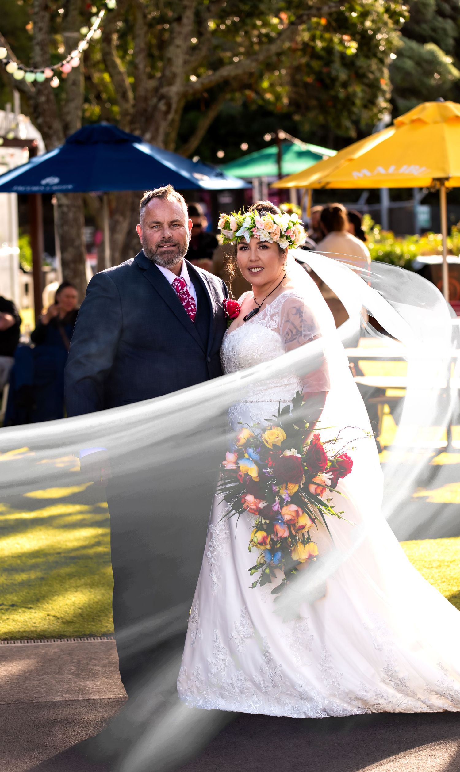 A wedding couple poses outdoors with the bride's veil flowing dramatically in the breeze during their outdoor celebration.