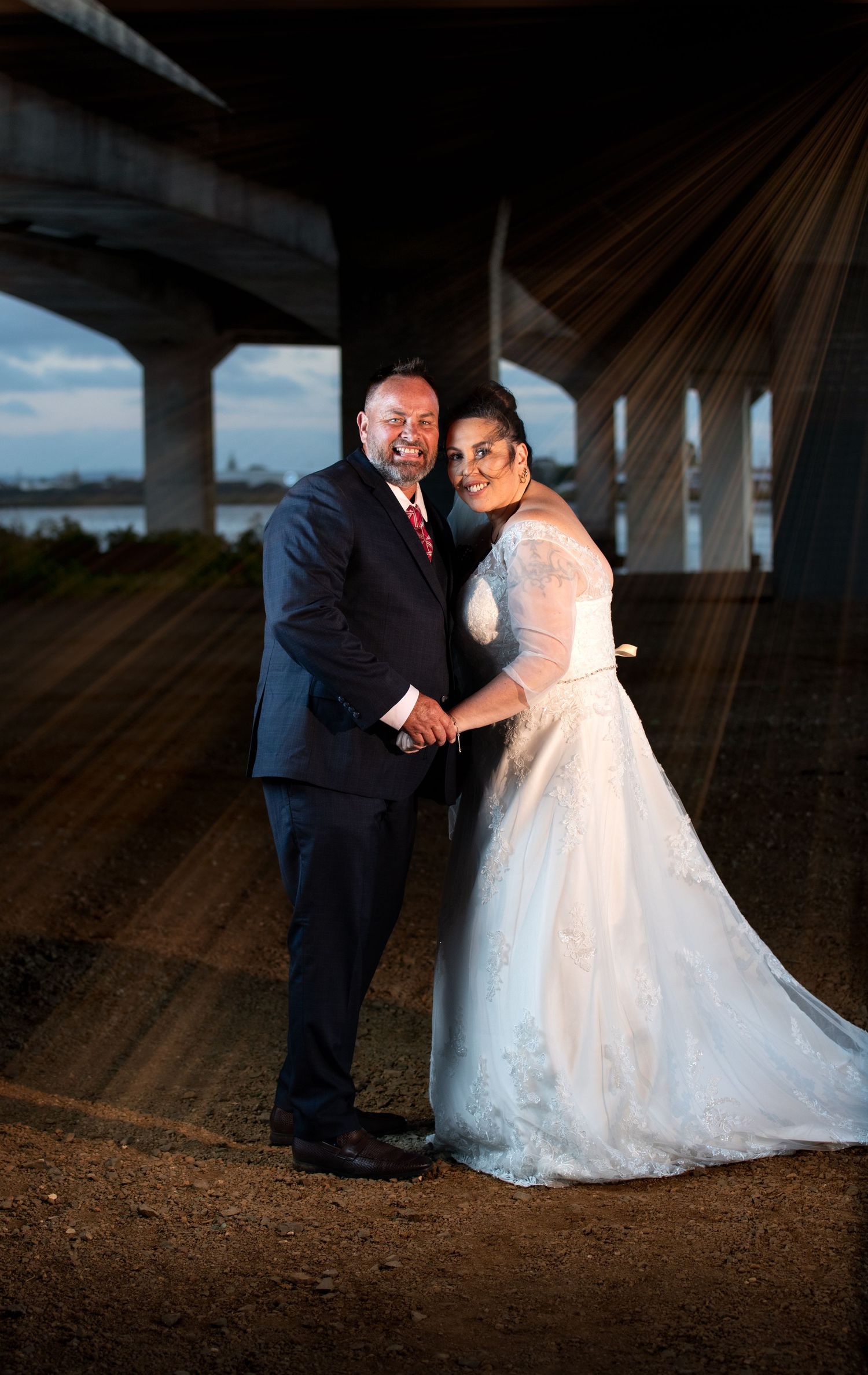 A couple in wedding attire poses under a bridge at sunset creating dramatic silhouettes and lighting effects.