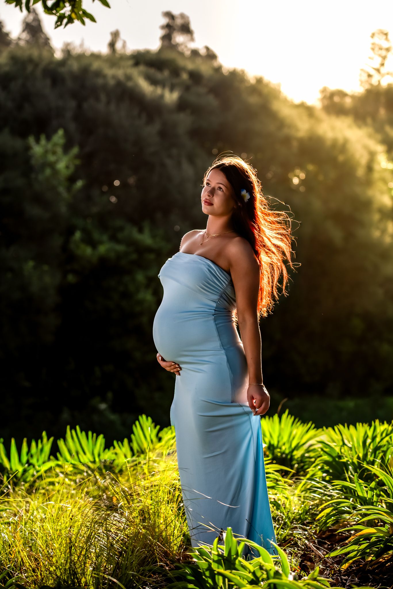 Pregnant woman in light blue dress standing in sunlit garden, cradling baby bump with long hair blowing in breeze.