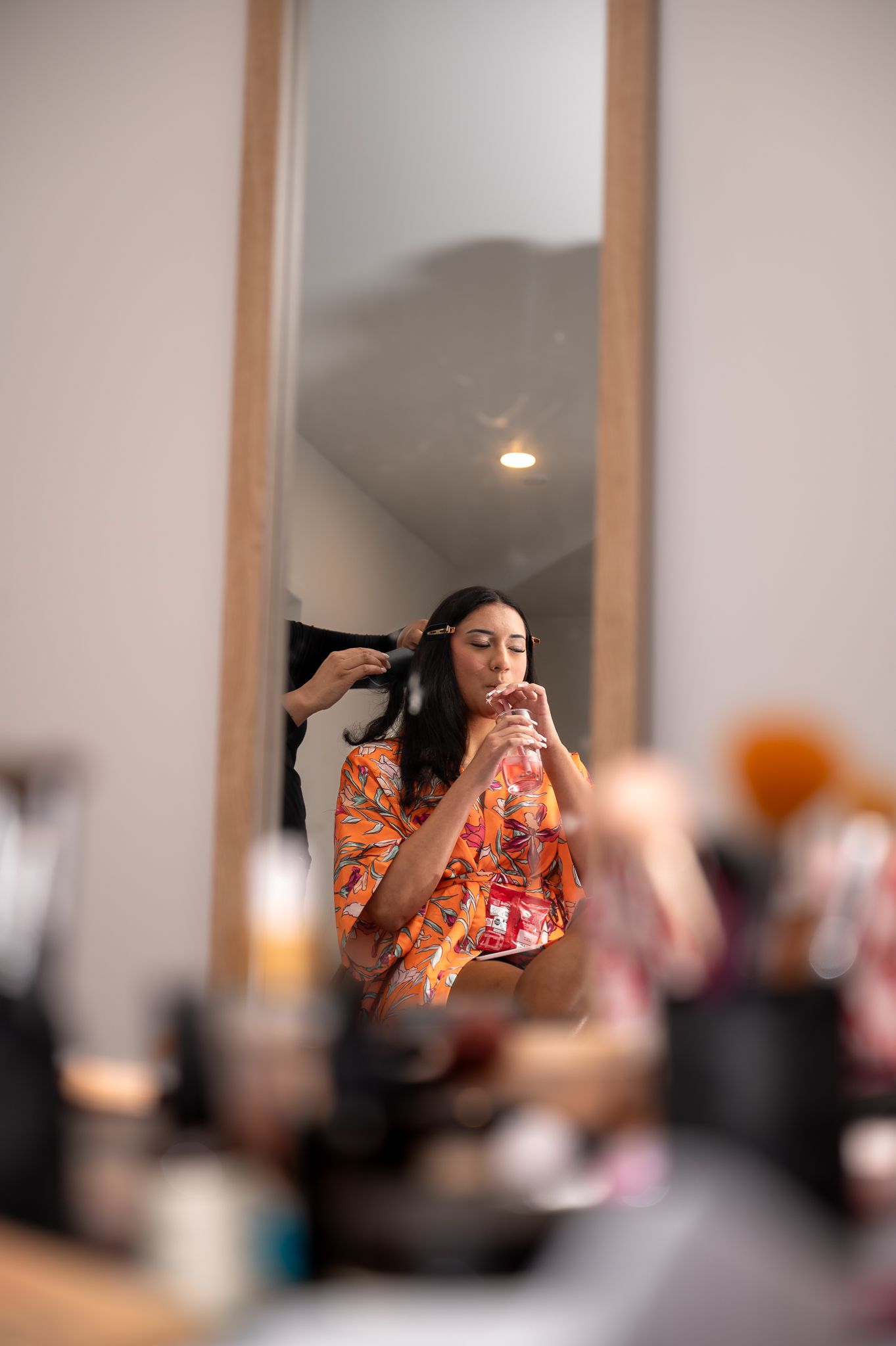 Woman in floral robe getting hair and makeup done, reflected in a wooden-framed vanity mirror.