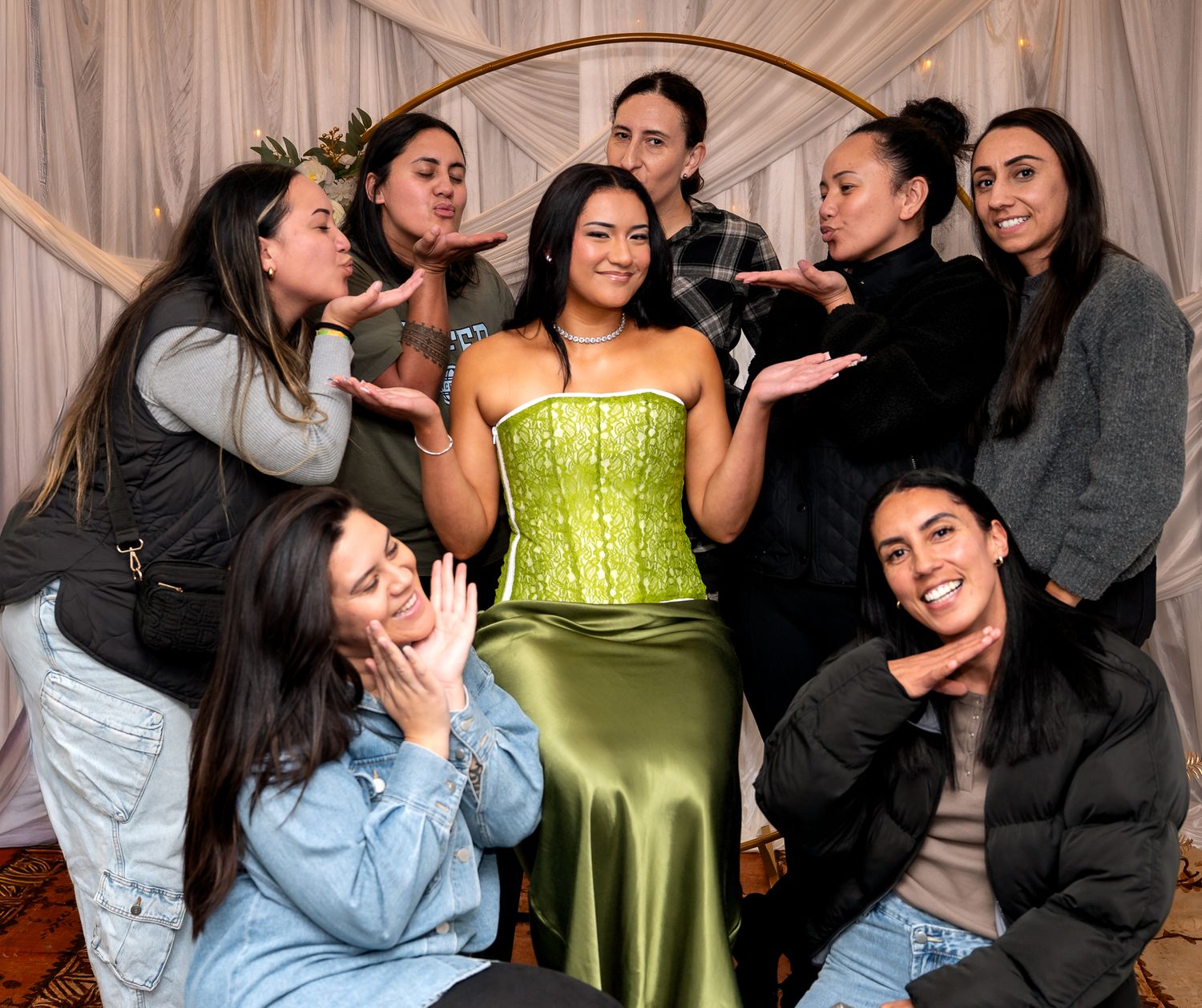 Group of women posing playfully around a woman in a green satin dress at a celebration event.