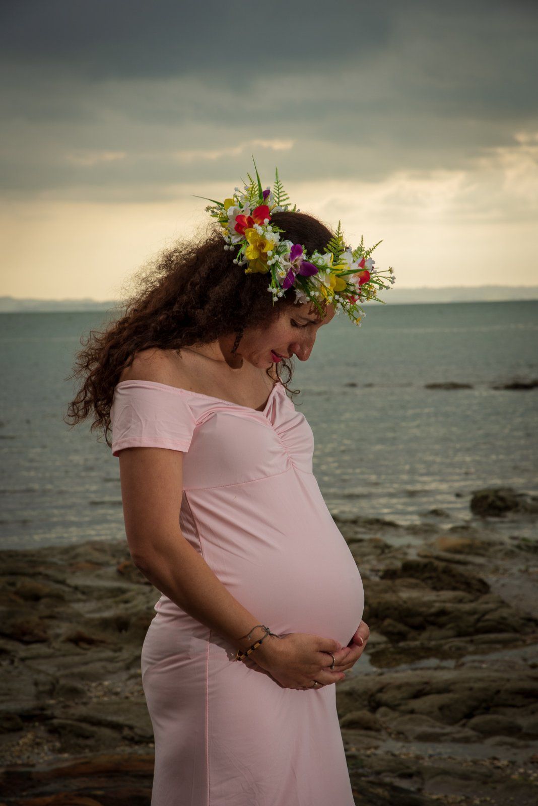 A pregnant woman in pink dress with flower crown poses against ocean sunset backdrop.