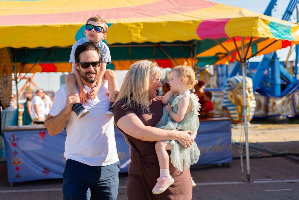 A group enjoys a sunny day at a carnival with colorful tents and vendor stands visible in the background.