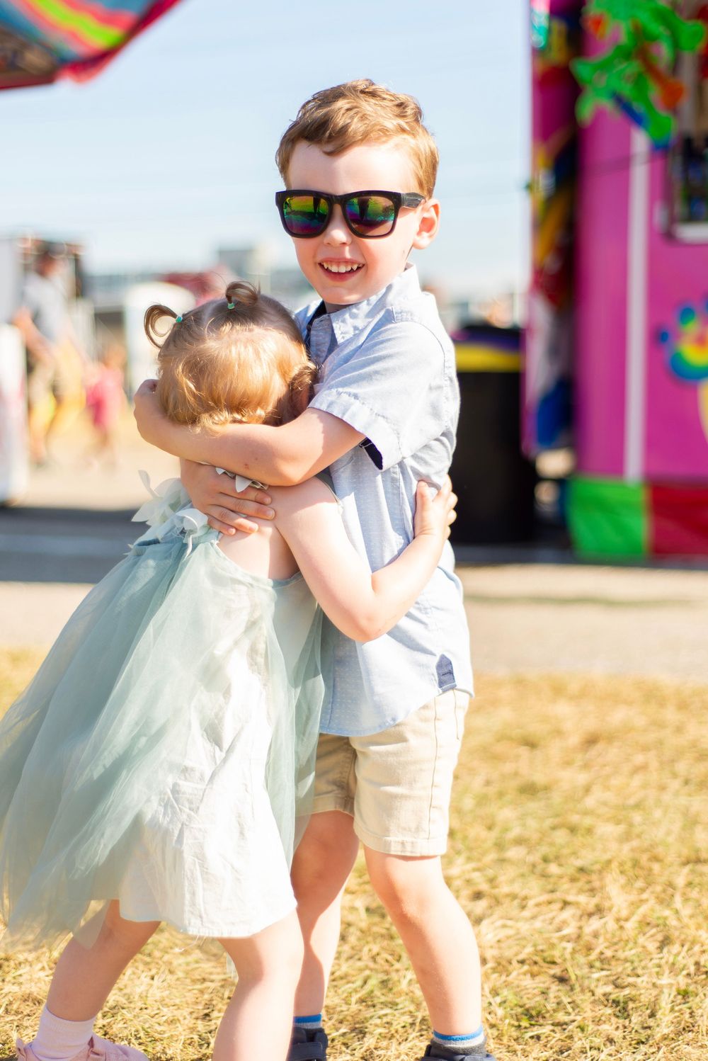Two children sharing a heartwarming hug at a festive carnival with colorful booths behind them.