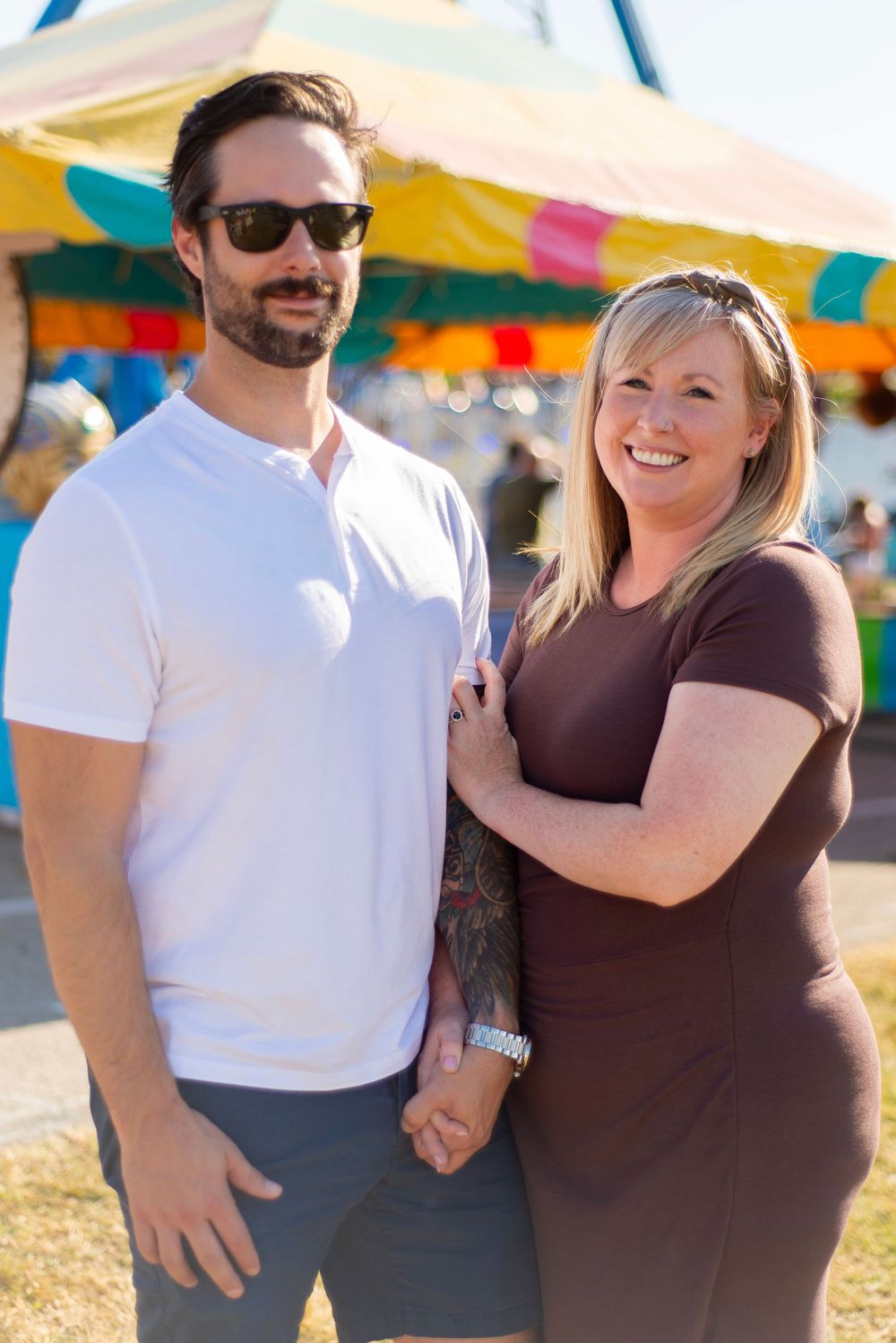 A couple stands outdoors at a festival with colorful tents in the background on a sunny day.