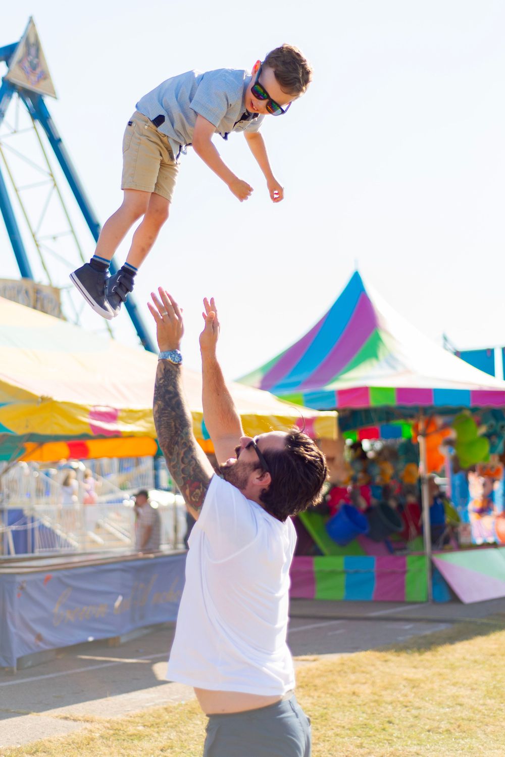 A playful moment at a carnival as someone lifts a child up in the air with colorful tents in the background.
