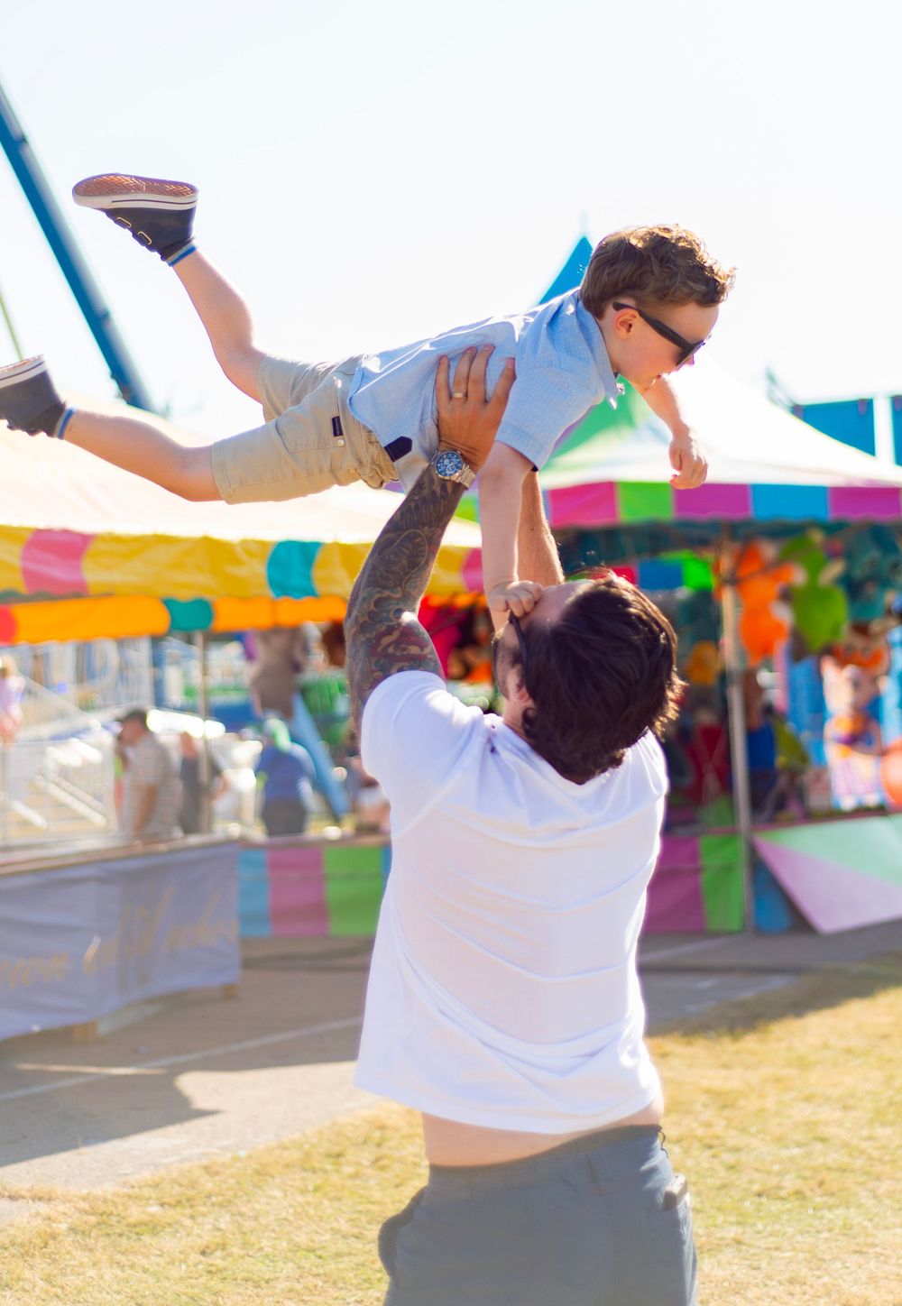 Playful moment at a carnival as an adult lifts a child high into the air against a backdrop of festival tents.