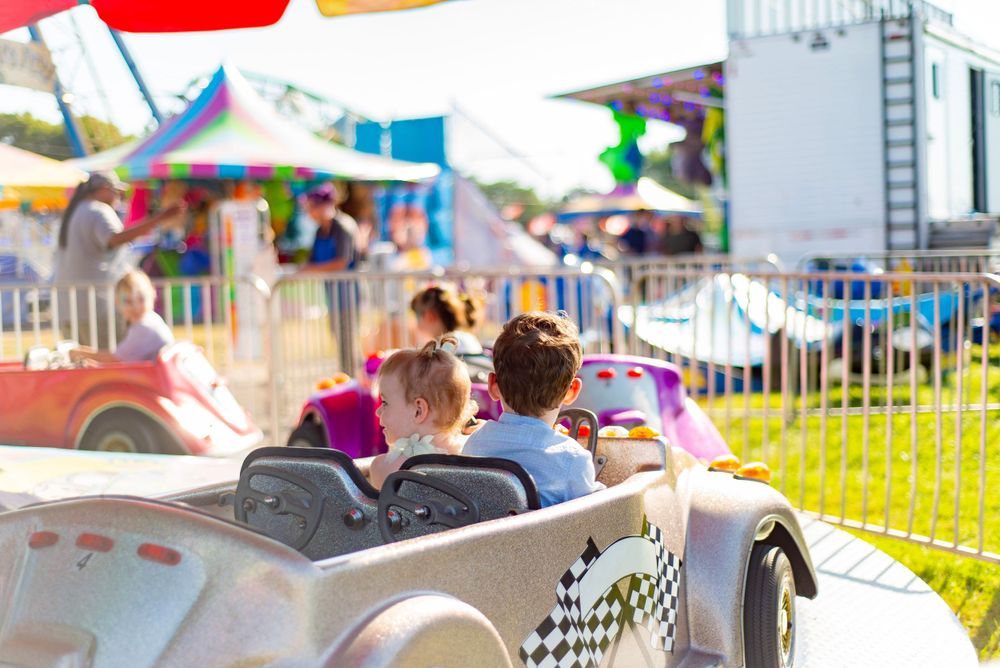 Children ride together in a silver car attraction at a colorful carnival fairground with tents and rides in the background.