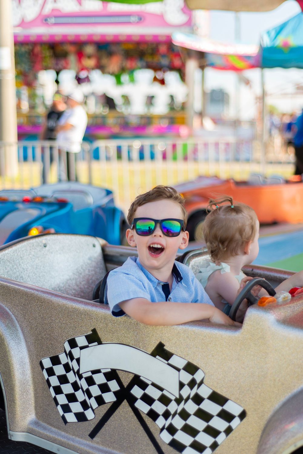 Child wearing blue sunglasses smiling with excitement while riding a carnival car attraction.