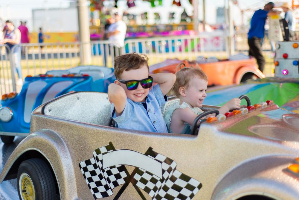 Two kids on a carnival ride smile and enjoy a race car attraction with checkered flag decor at an amusement park.