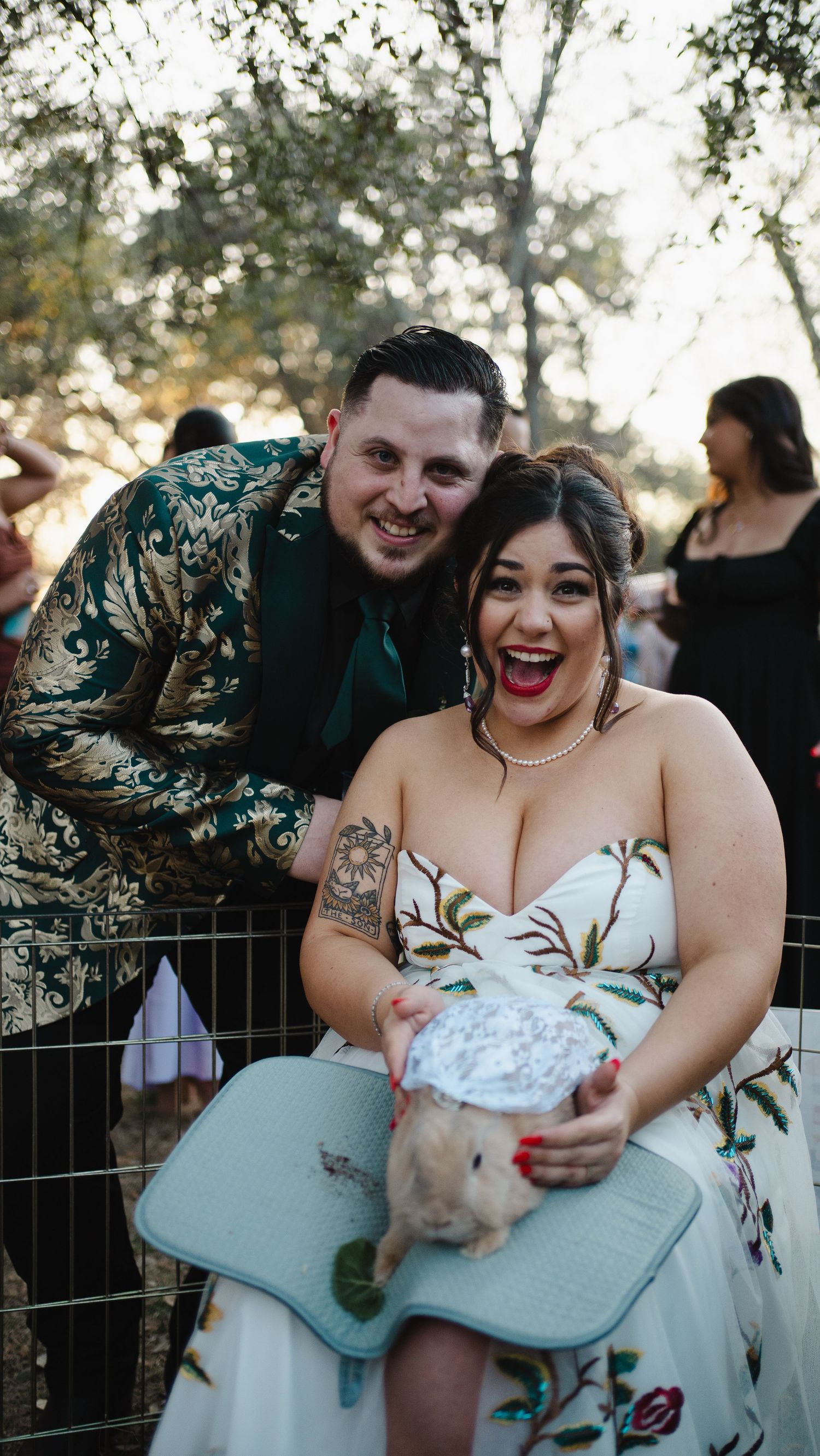 A happy couple at an outdoor event poses with a small rabbit while wearing elegant floral and patterned formal attire.