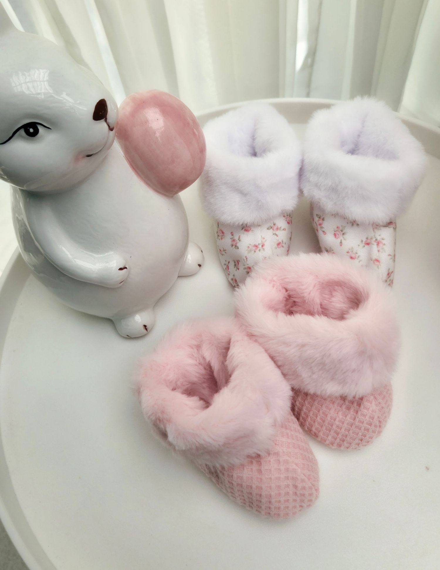 Two pairs of pink fluffy baby booties with floral accents displayed beside a white ceramic bunny figurine.