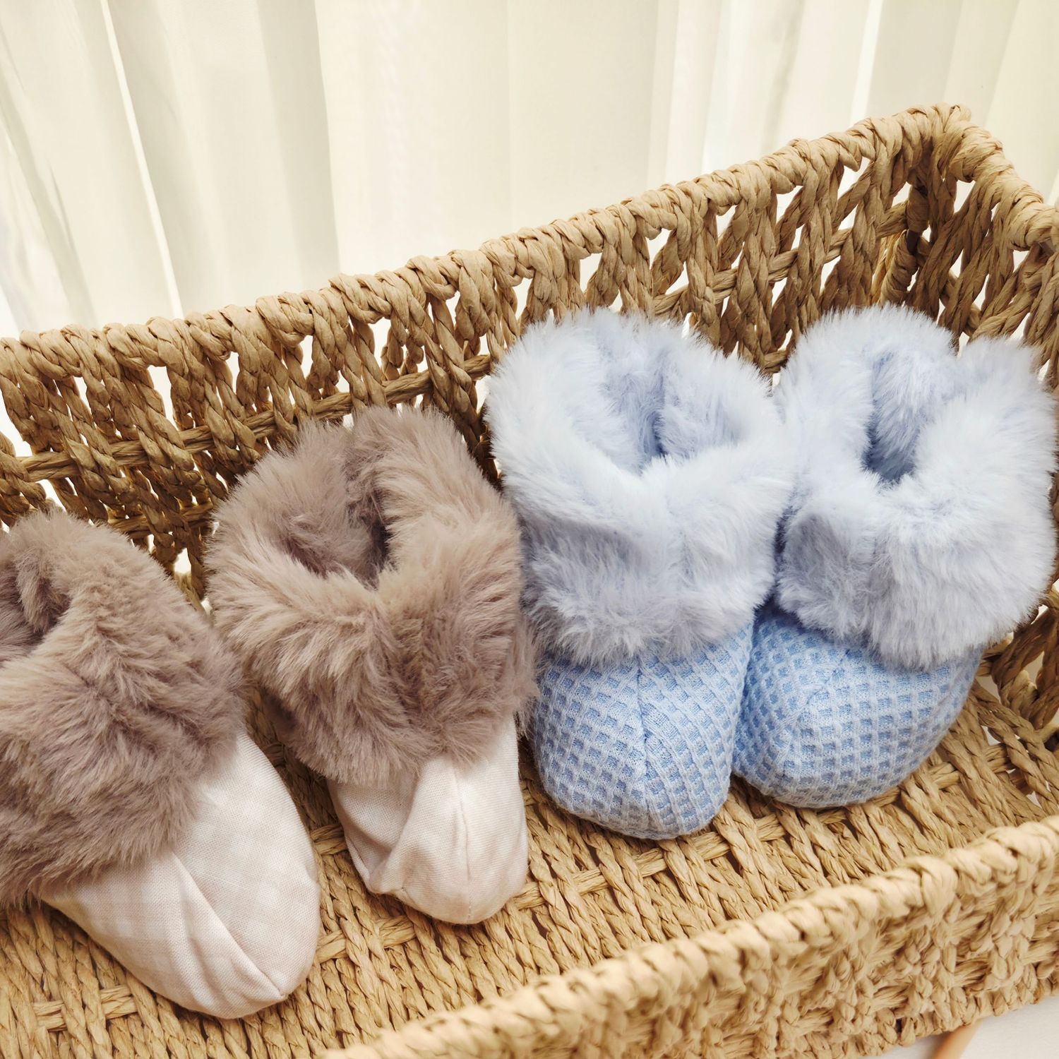 Two pairs of fluffy baby booties, one gray and one blue, displayed in a woven basket.