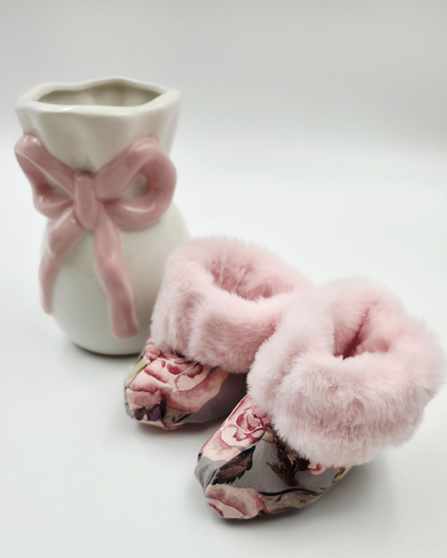 Pink floral baby booties with faux fur trim beside a white ceramic vase with pink bow ribbon decoration.