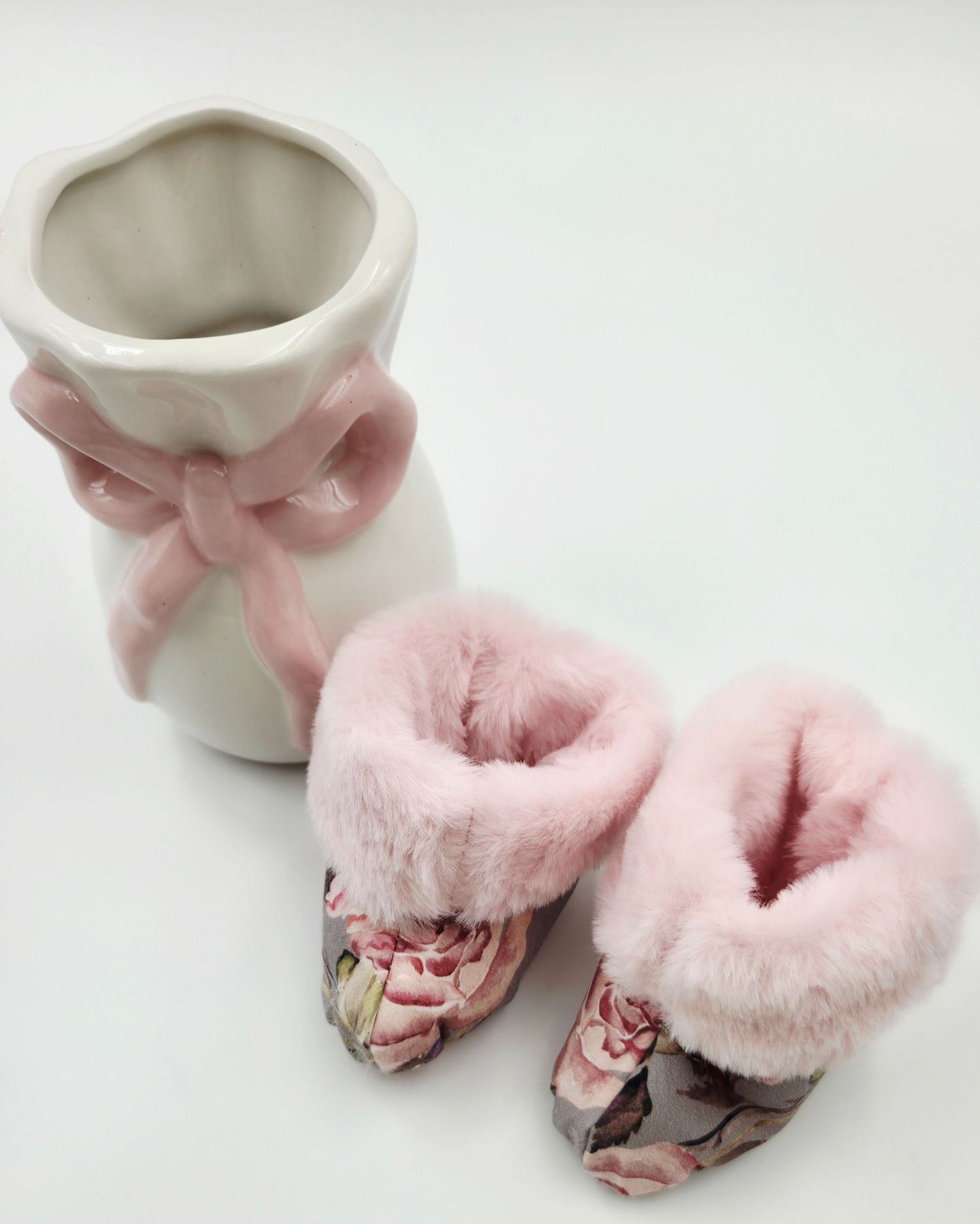 Pink floral baby booties with faux fur trim beside a white ceramic vase with pink ribbon bow.