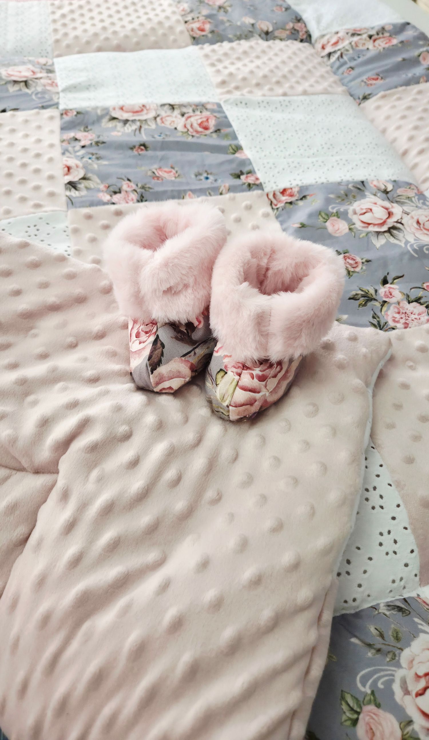 Pink floral baby booties with faux fur trim resting on a soft pink minky dot blanket with a matching rose patchwork quilt.
