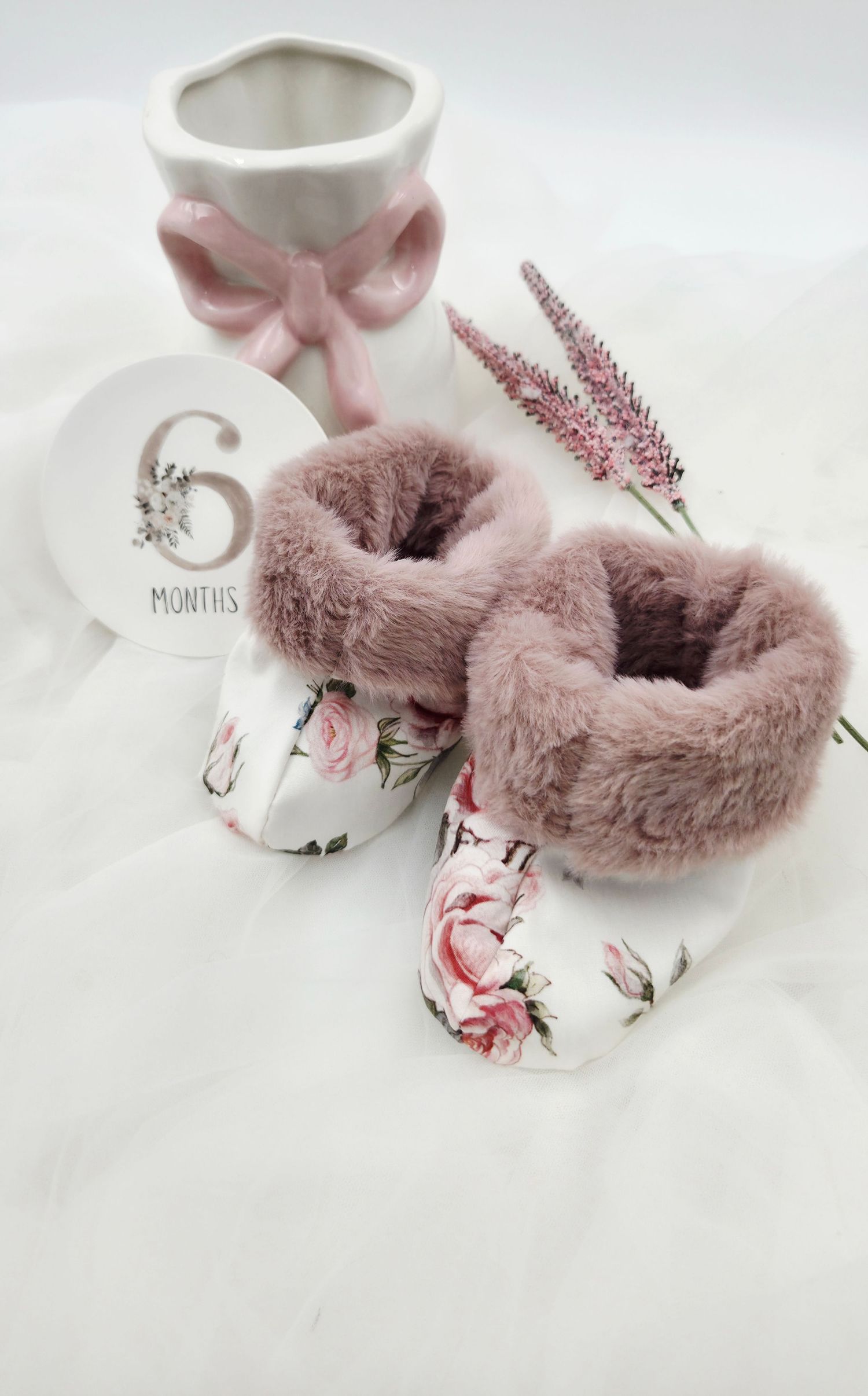 Floral baby booties with pink fur trim beside a 6 months milestone disc and ceramic bow vase.